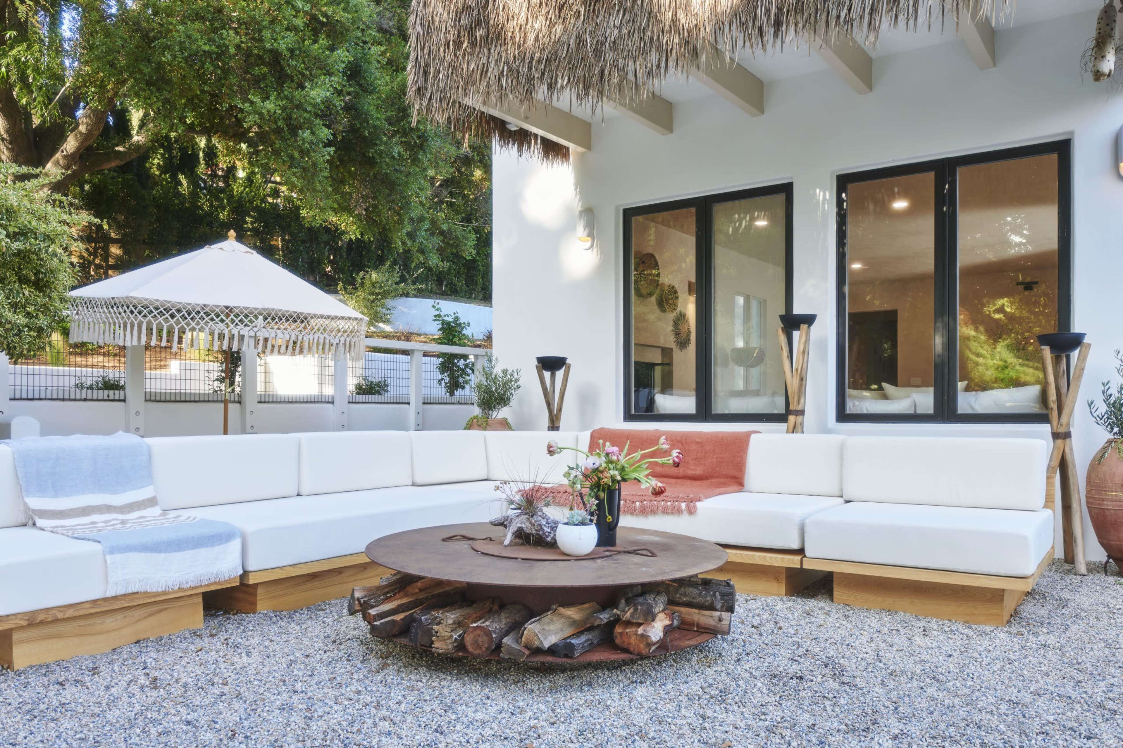 A modern outdoor seating area features a circular wooden coffee table surrounded by white cushioned sofas and decorative elements under a thatched roof.