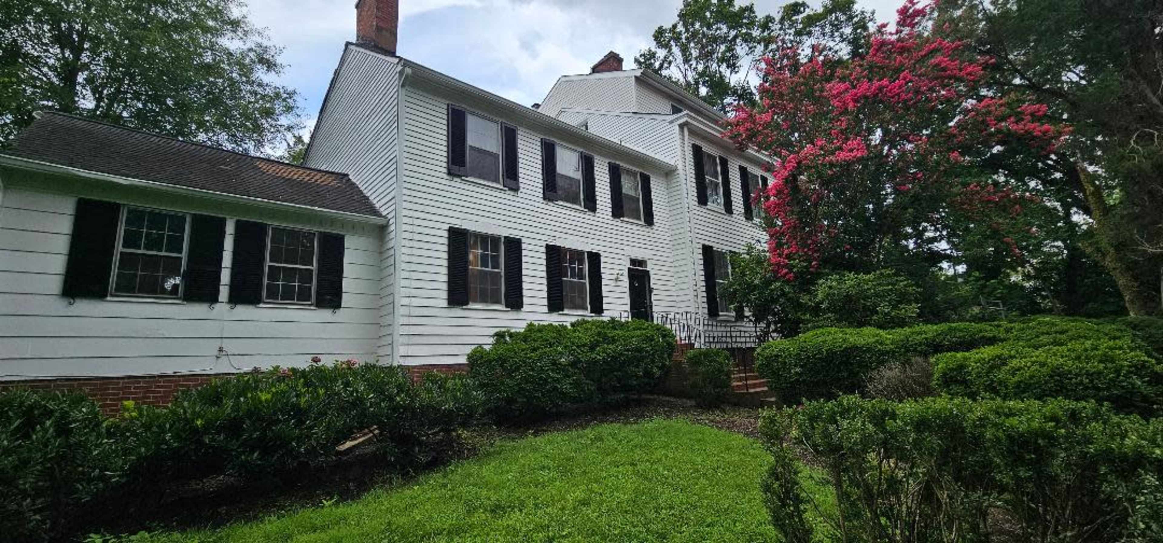 A white two-story house with black shutters sits beside a lush green landscape and flowering bushes.