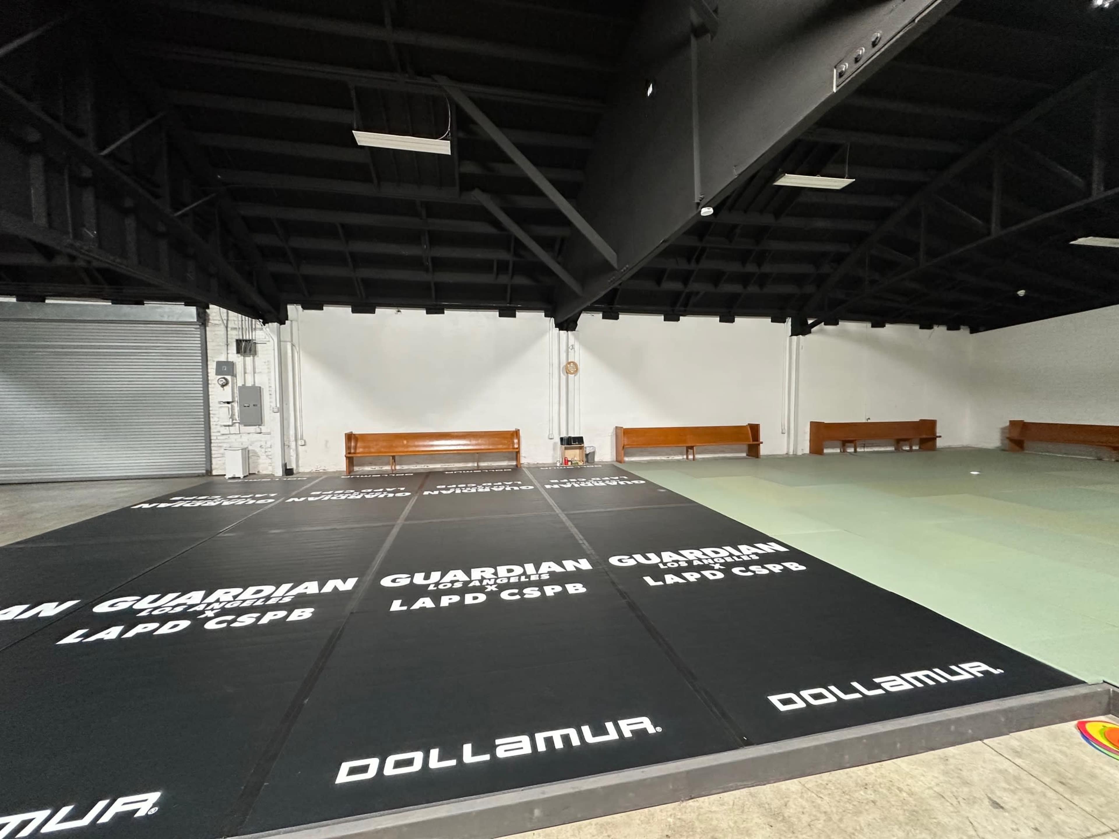 The image shows a gymnasium area with black mats featuring logos, surrounded by benches and a large open space under a black ceiling.