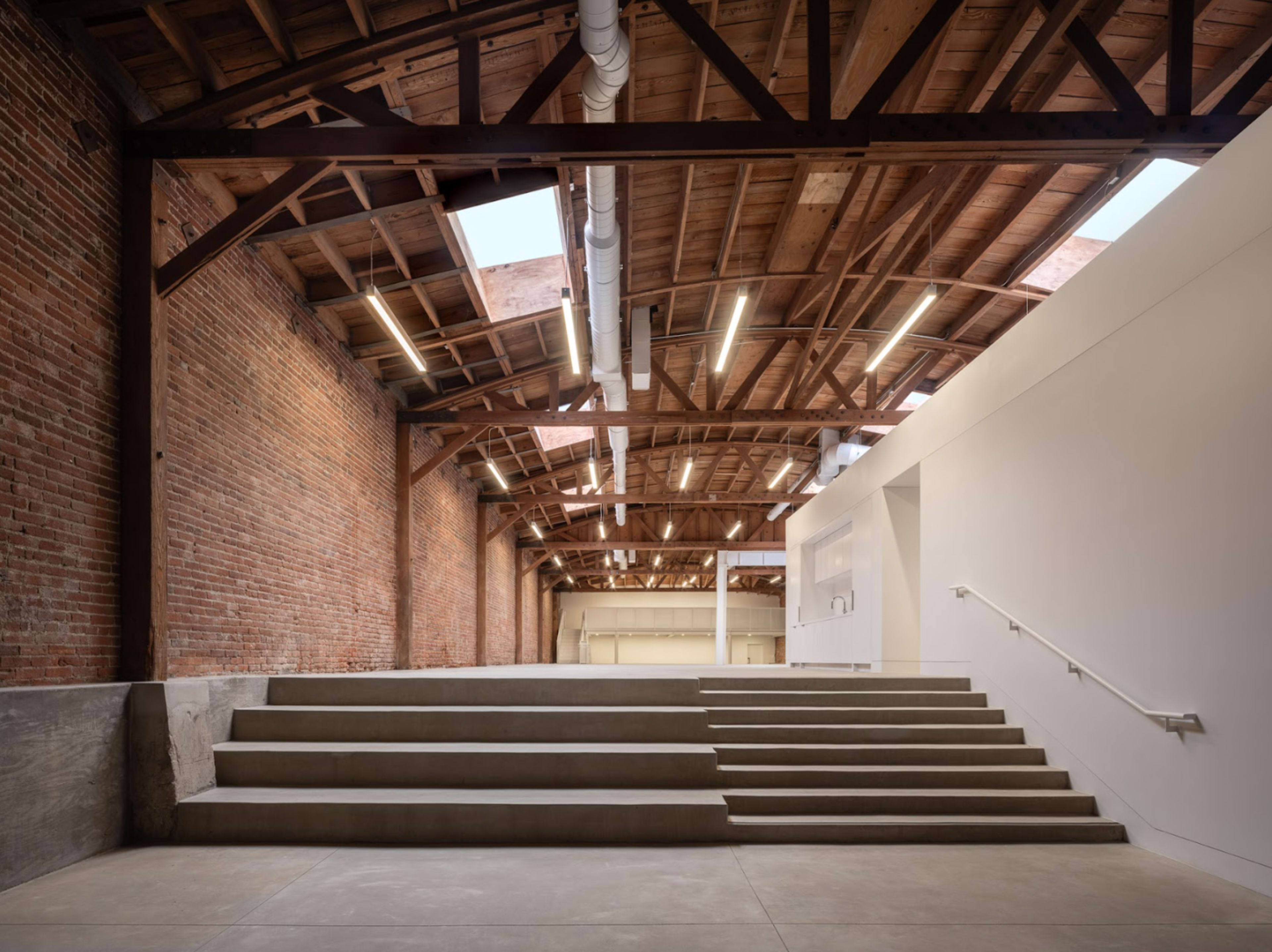 A spacious interior with exposed brick walls, wooden beams, and a concrete staircase leading to a white wall that separates two areas.