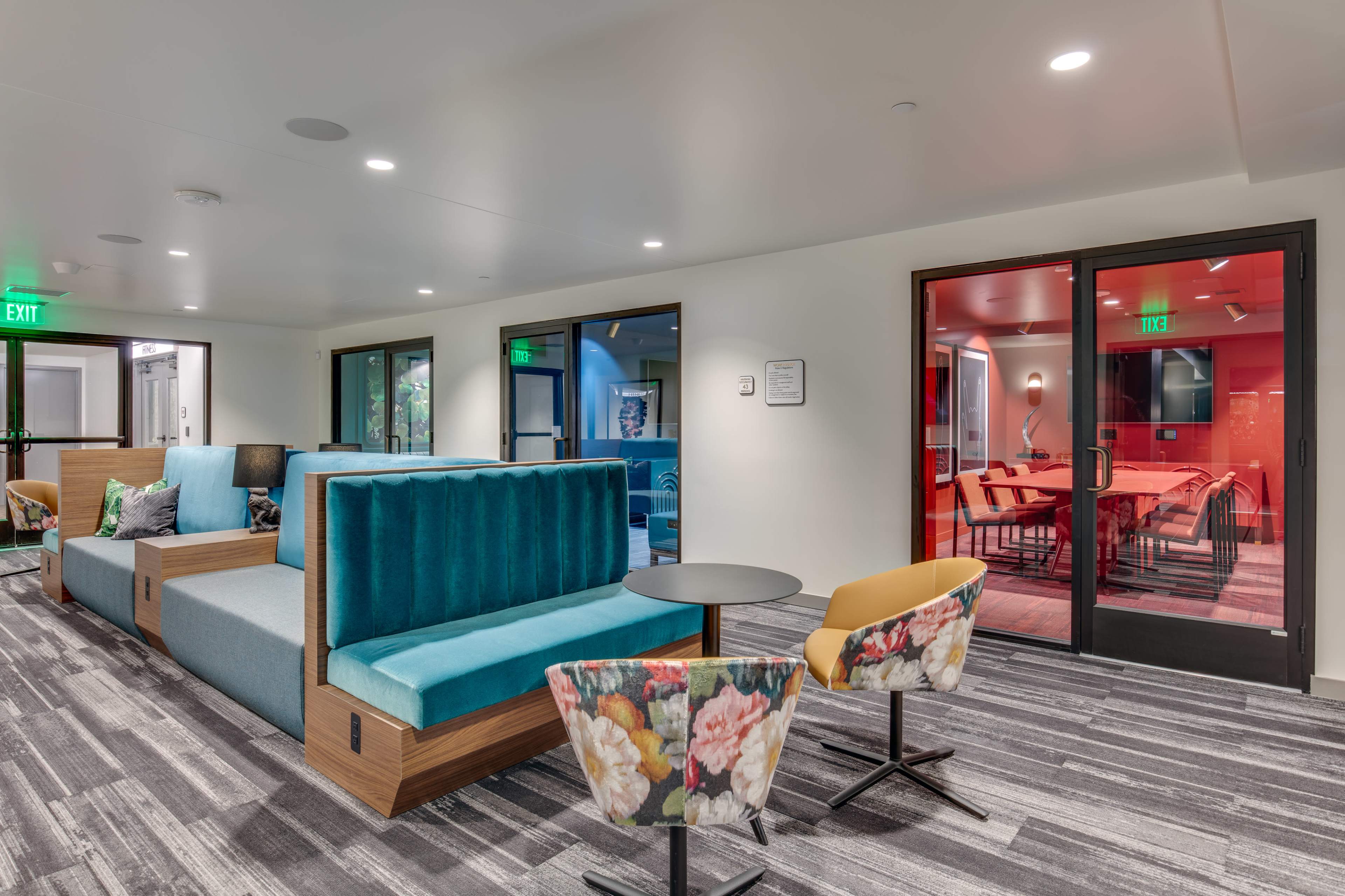 A modern lounge area with teal seating, floral chairs, and glass doors leading to a red-lit conference room.