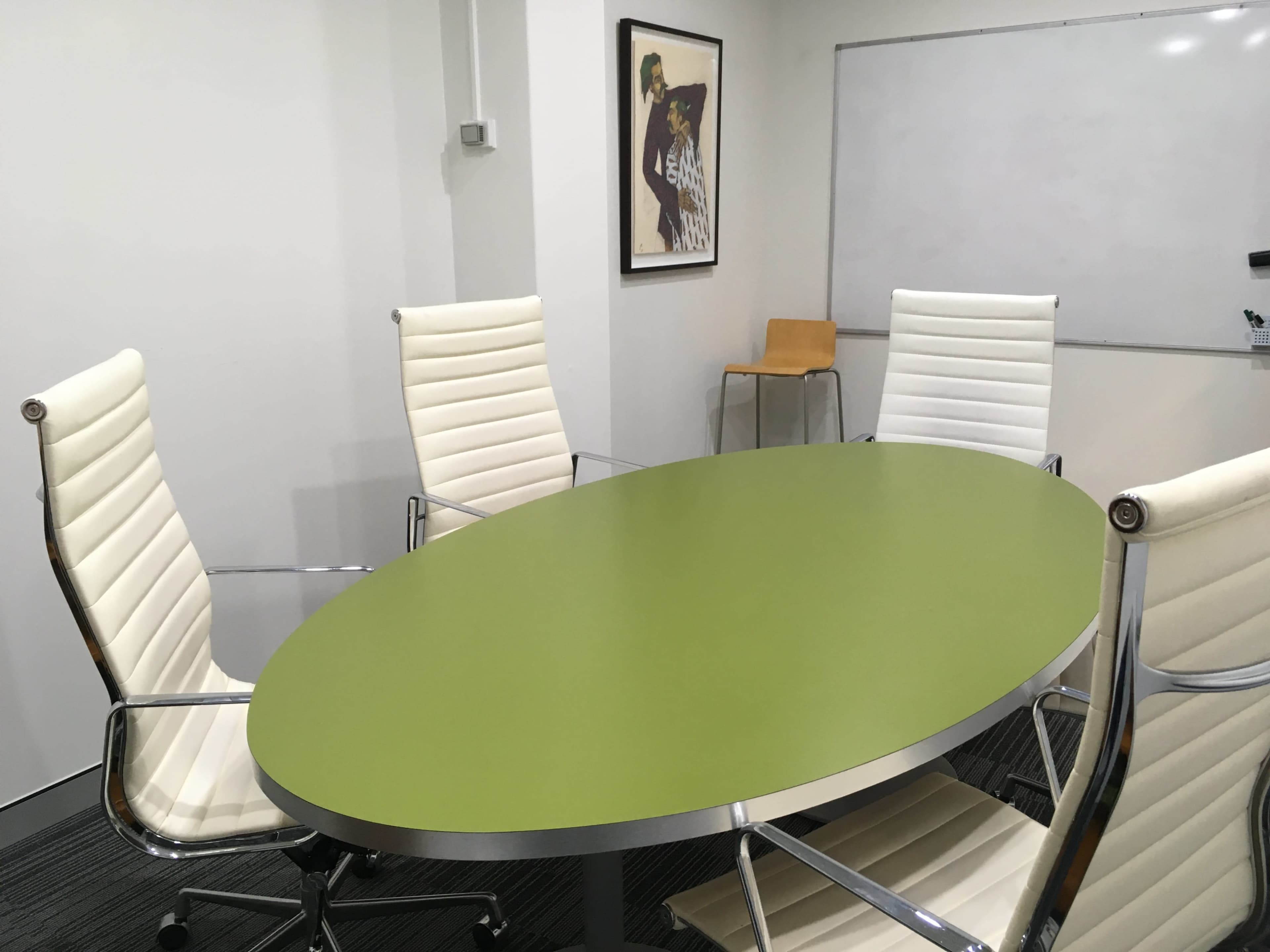A modern conference room features a green oval table surrounded by white leather chairs, with a framed artwork on the wall and a whiteboard in the background.