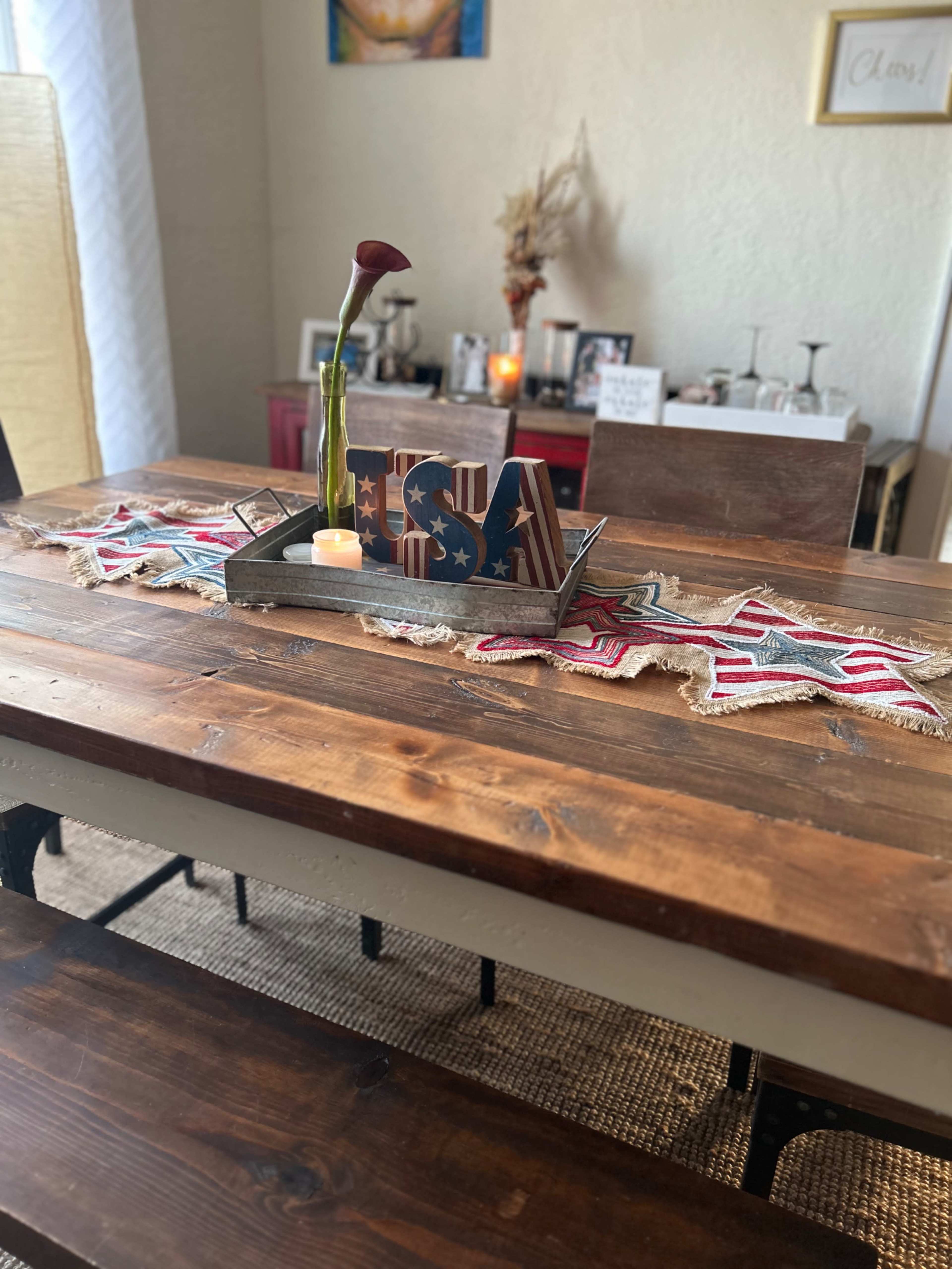 A wooden dining table is adorned with a patriotic decoration featuring the letters "USA," a small candle, and a vase, with a rustic table runner underneath.