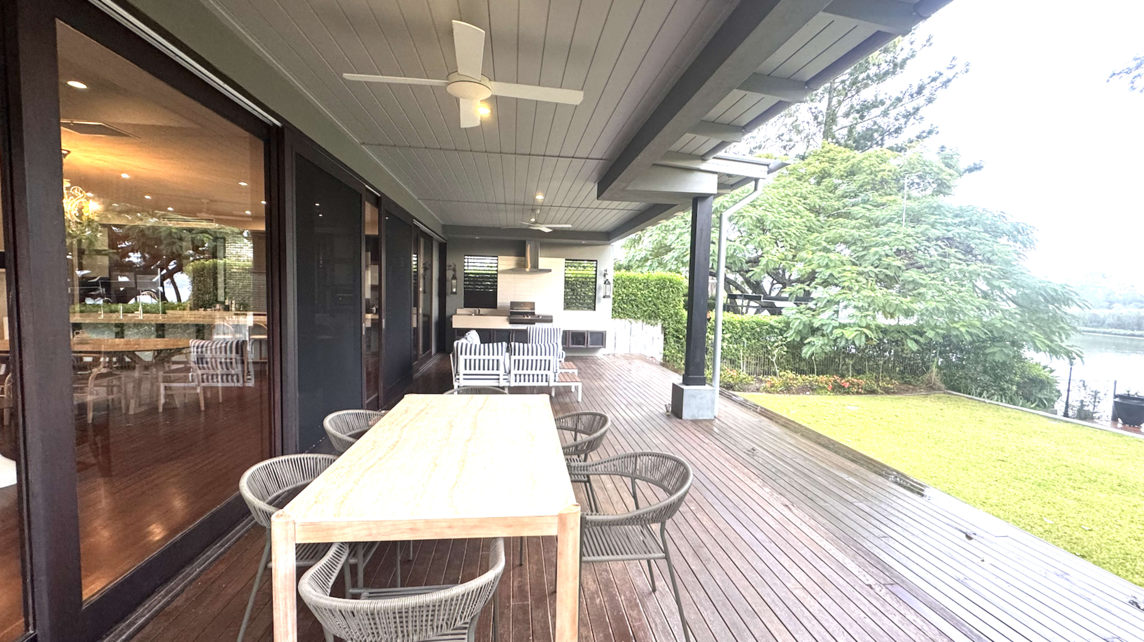 The image shows a modern outdoor patio with a wooden table and chairs, overlooking a grassy area and water beyond.