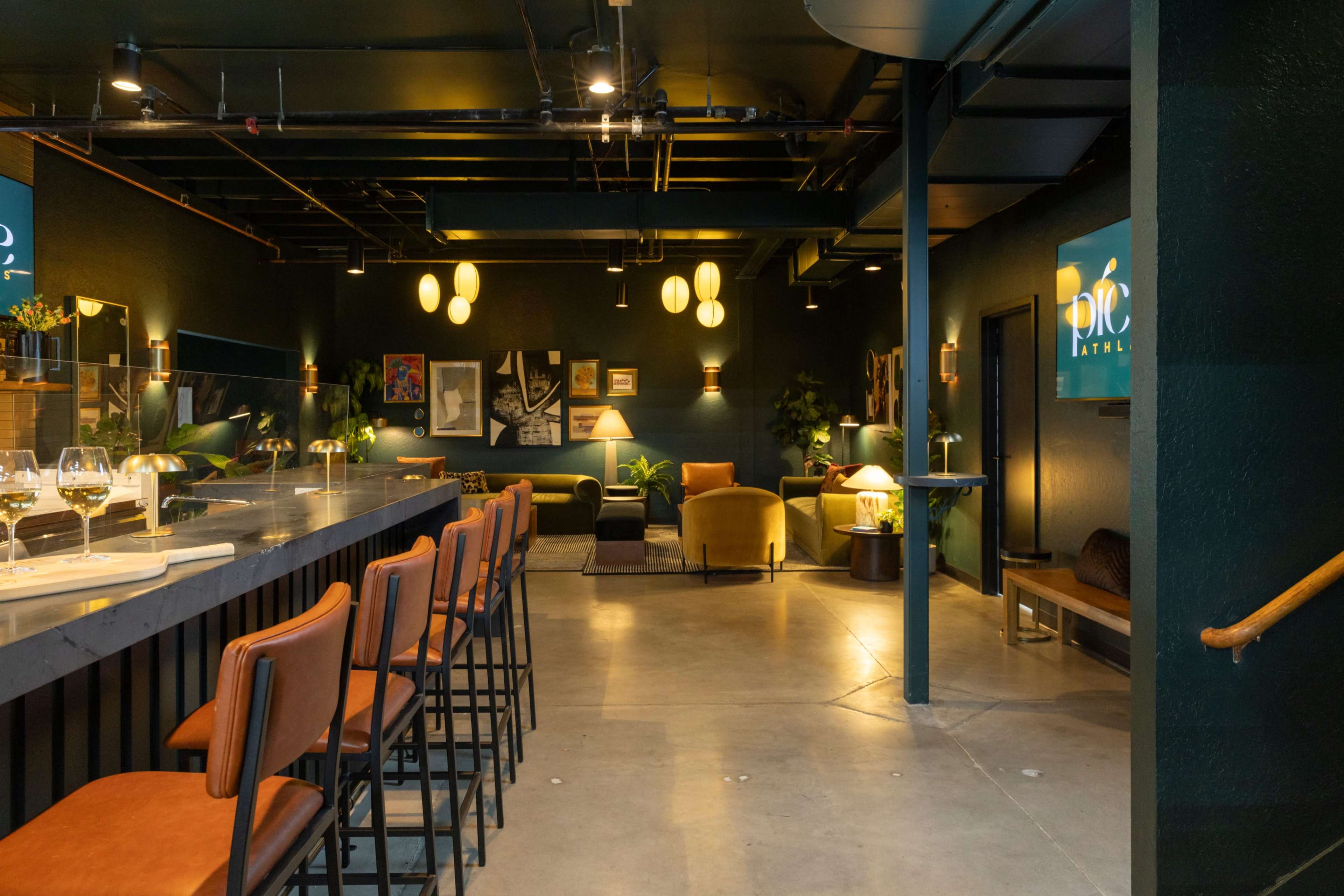 The image shows a modern bar area with high stools lined up against a dark countertop, featuring seating areas adorned with artwork and decorative lamps.