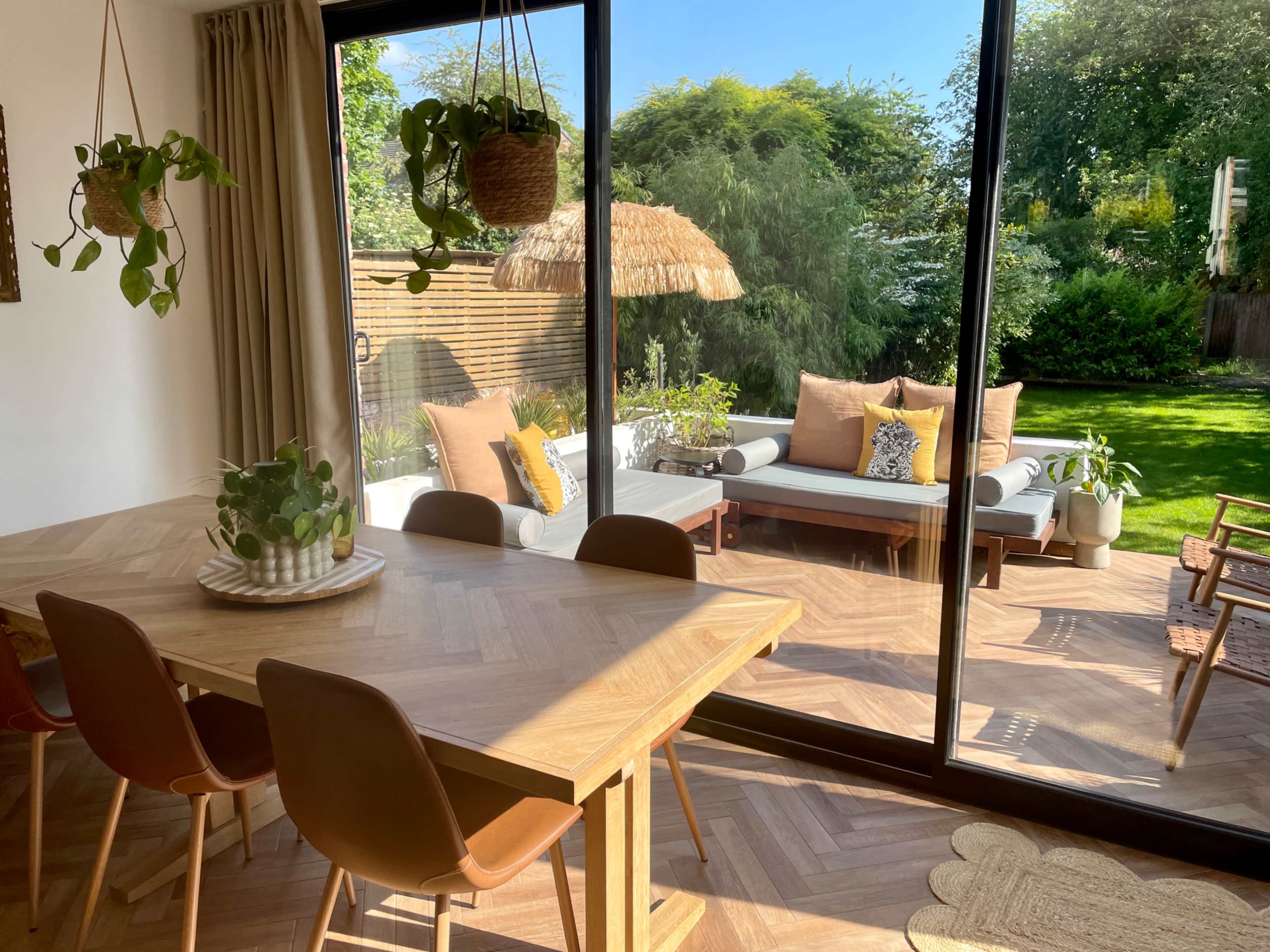 The image shows a modern dining area with a wooden table and chairs, overlooking a garden through large glass doors.