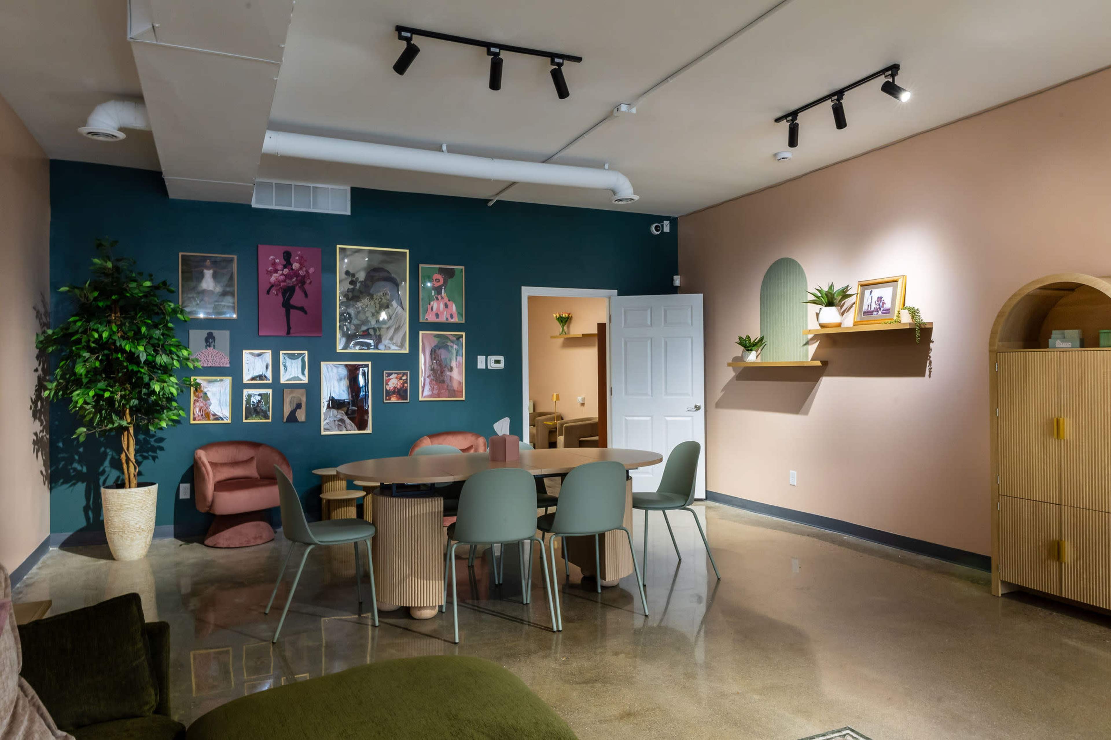 The image shows a modern, inviting interior space featuring a table surrounded by chairs, a gallery wall with framed art, and plants arranged in decorative pots.