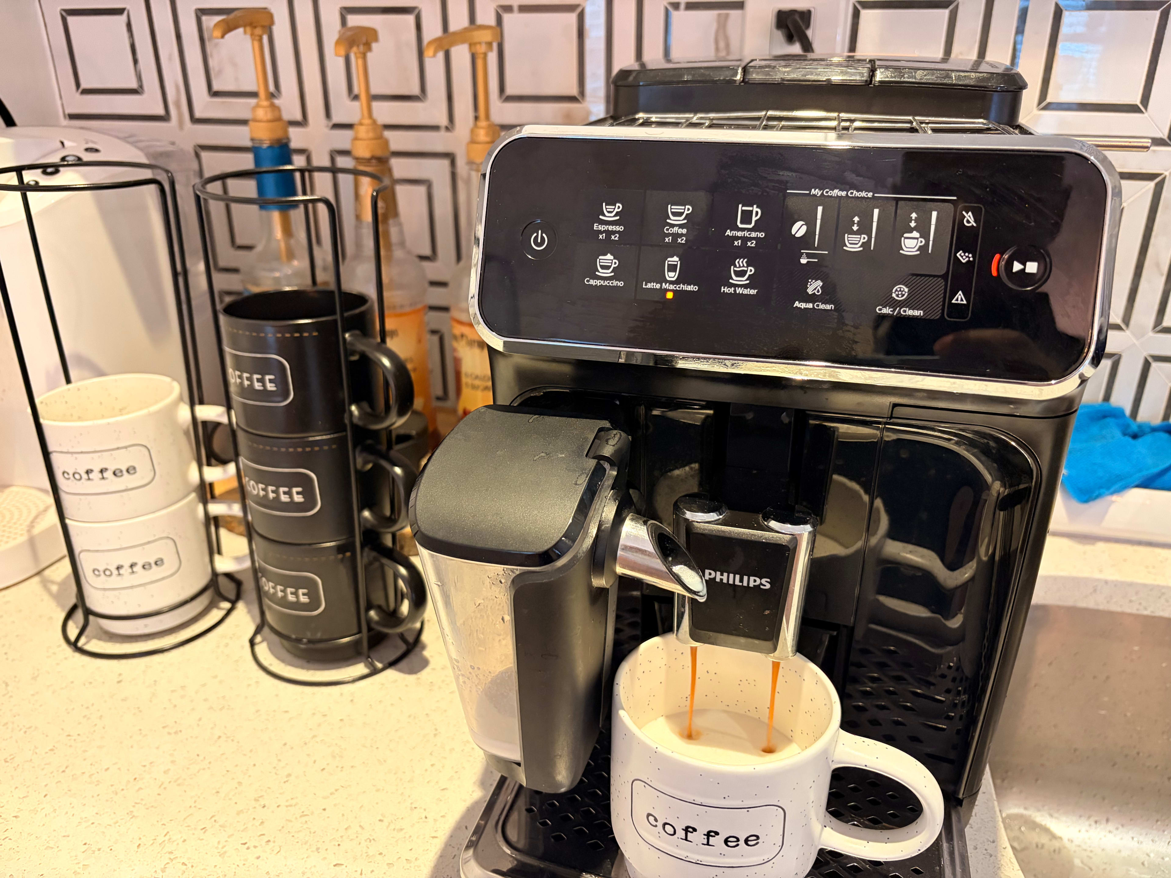 A coffee machine dispenses coffee into a white cup labeled "coffee," with additional cups and tools visible on the counter.