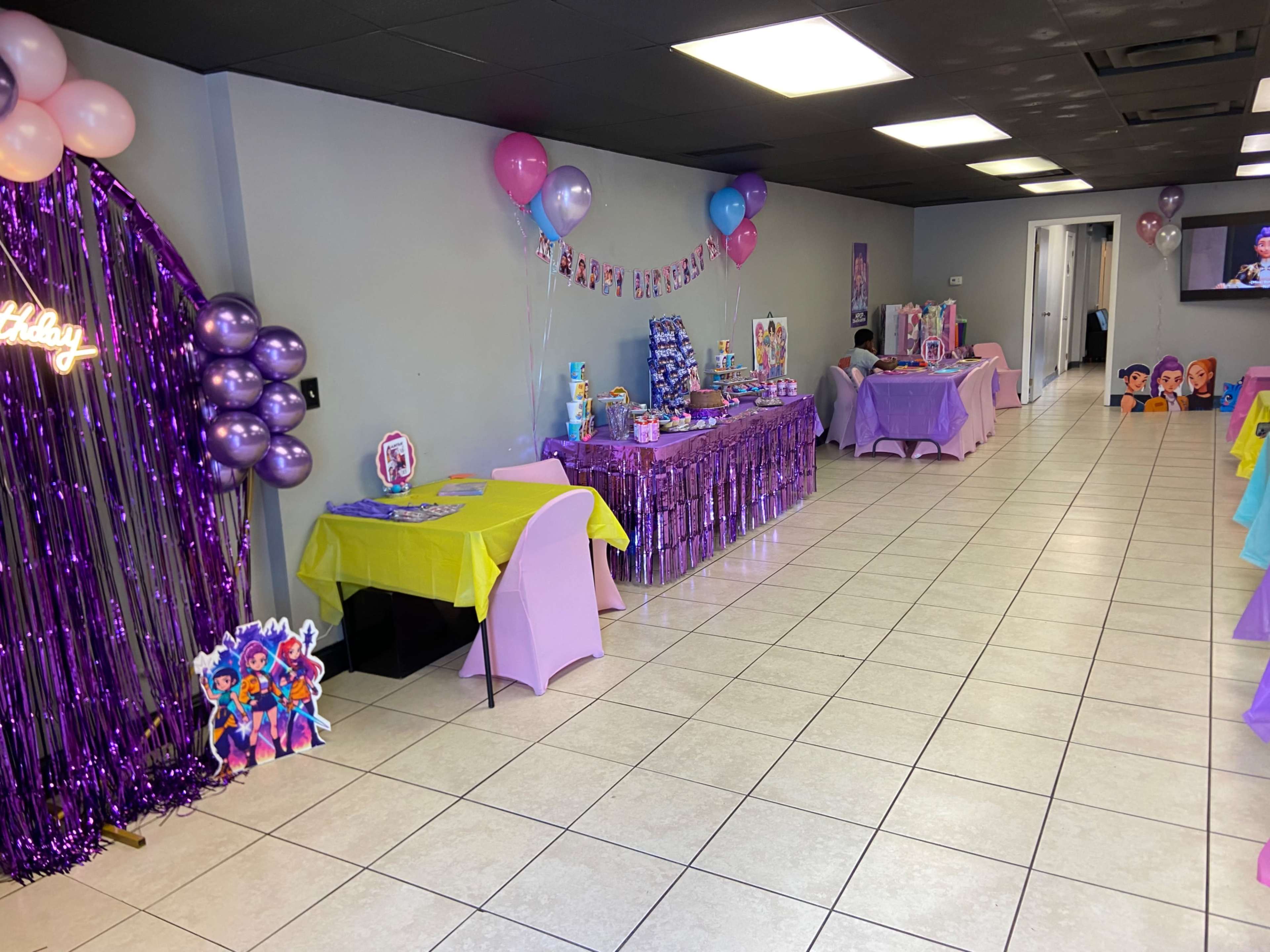 The image shows a decorated room set up for a birthday party, with multiple tables draped in yellow and purple tablecloths, balloons, and various party supplies displayed on the tables.