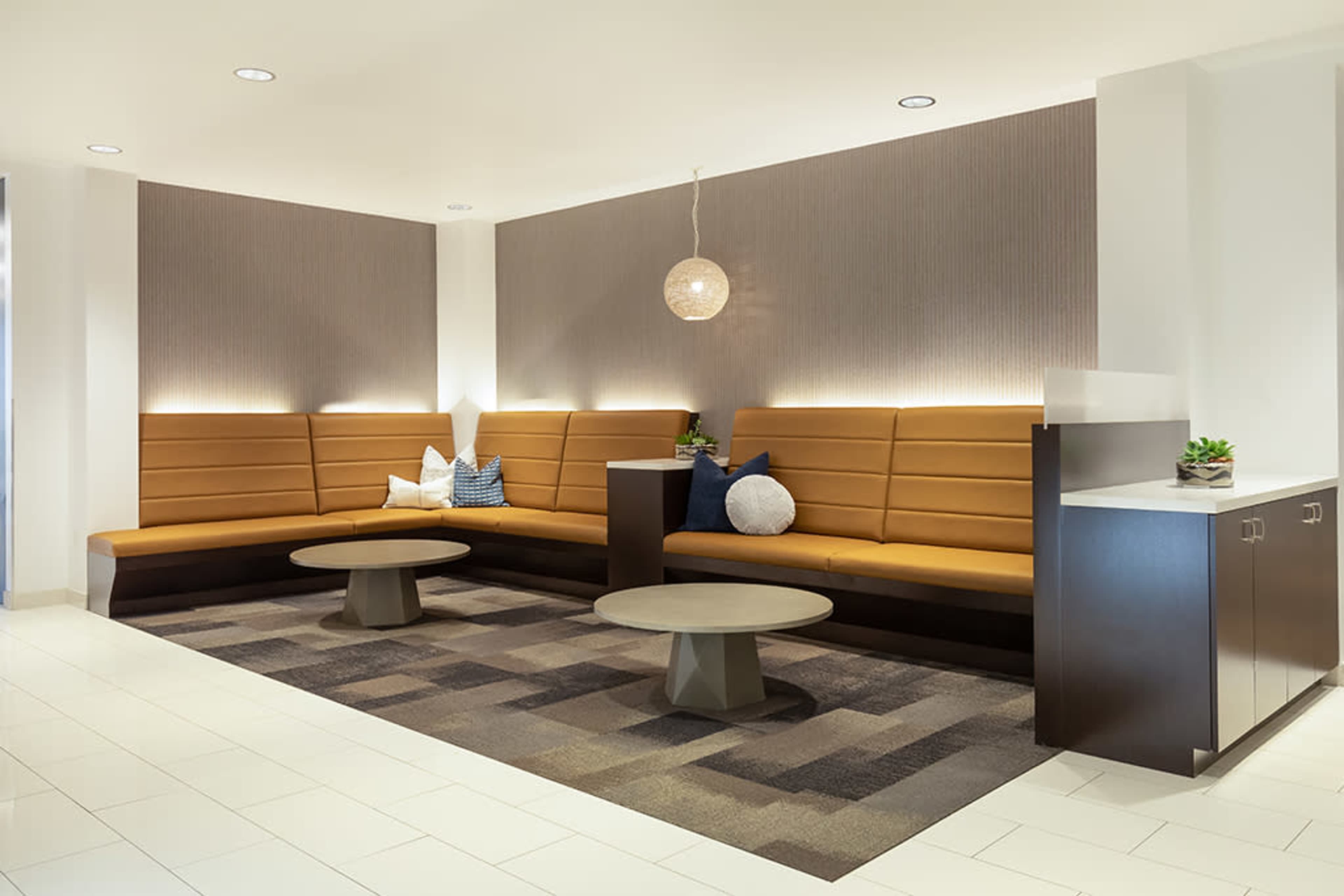 The image shows a modern waiting area with a long brown seating bench, two round coffee tables, and decorative pillows against a light-colored wall.
