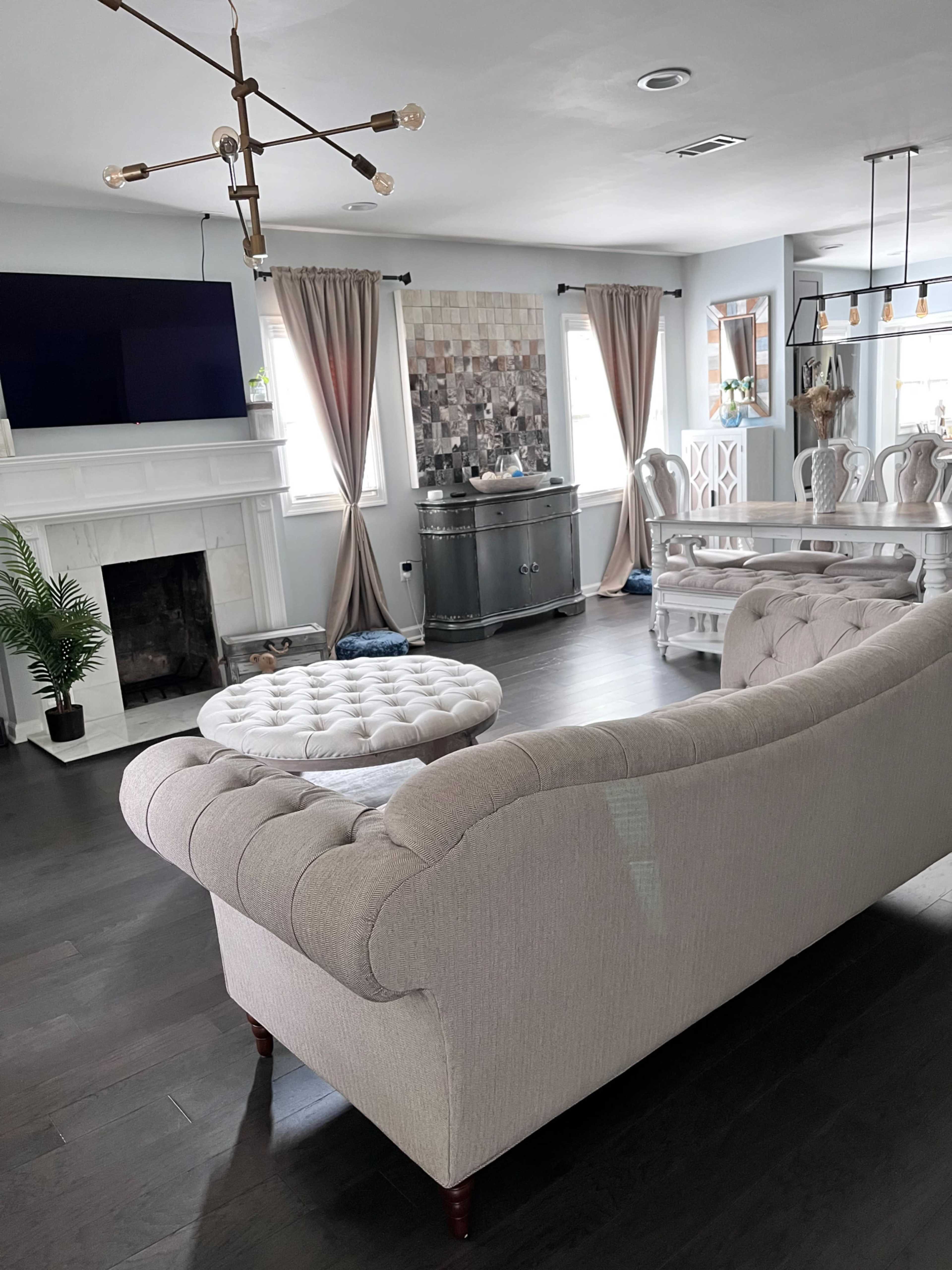 The image shows a modern living room with a light-colored tufted sofa, a round coffee table, a TV mounted on the wall, a dining area with a large table, and a decorative wall panel made of square tiles.