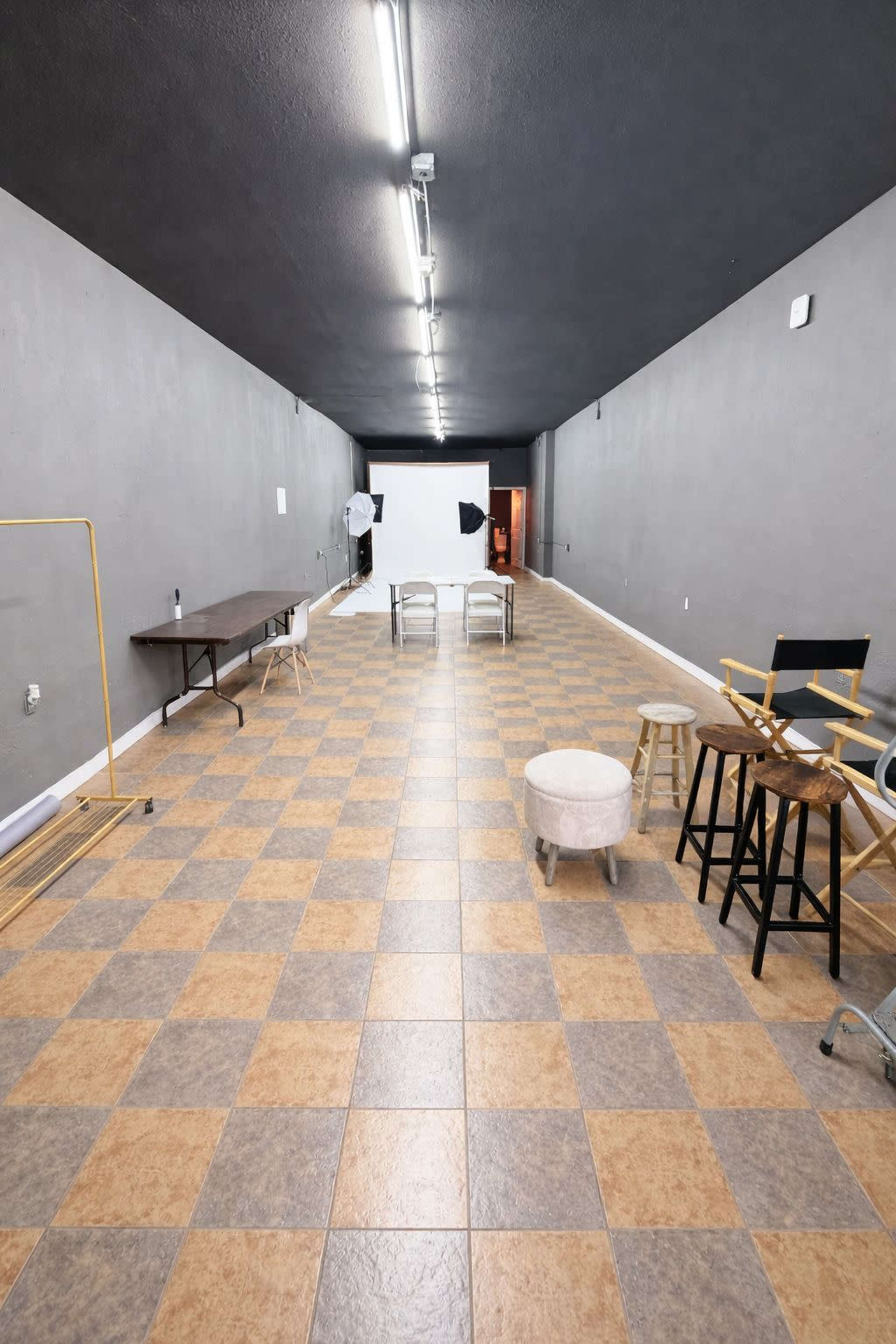 The image shows a spacious, empty studio with gray walls and a checkered floor, featuring a few chairs, tables, and lighting equipment set up for a photo shoot.
