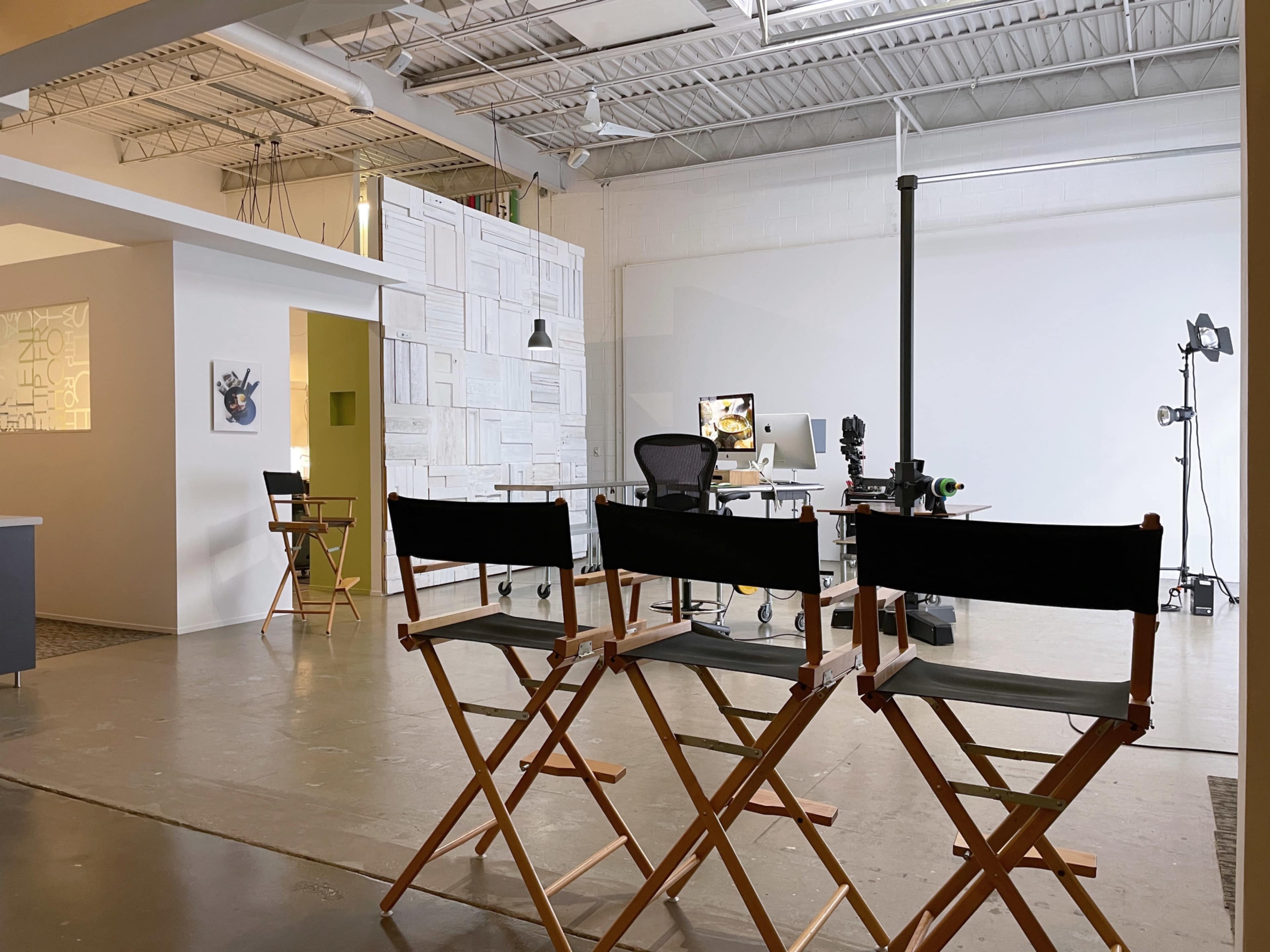 The image shows a spacious, well-lit studio featuring wooden director's chairs, a work desk with equipment, and a blank white backdrop.