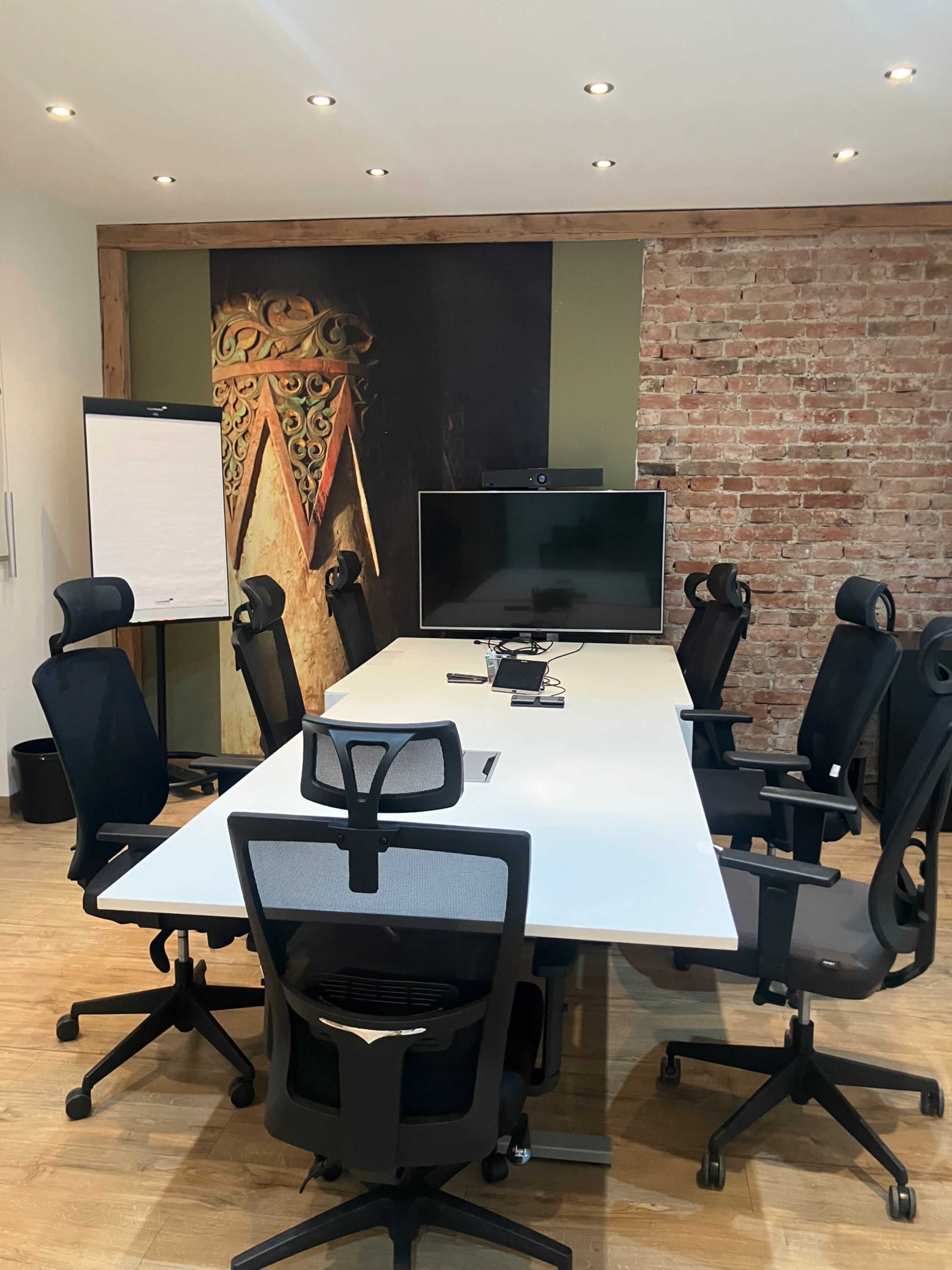 The image shows a modern conference room with a long white table, several ergonomic chairs, a wall with exposed brick, and a large screen mounted in front.