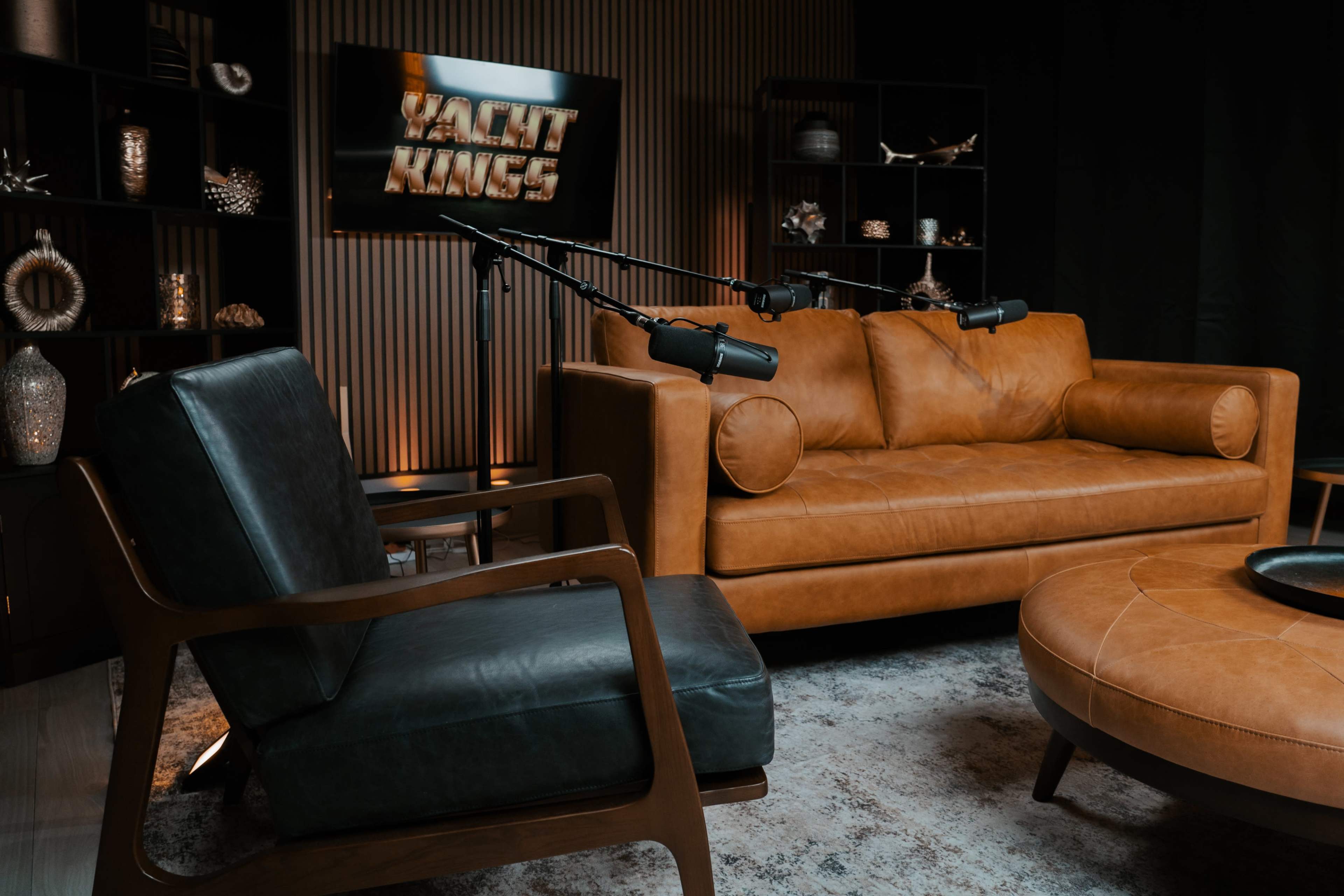 The image shows a stylish living room setup featuring a brown leather sofa, a black armchair, a round coffee table, and a wall-mounted screen displaying the text "Yacht Kings."