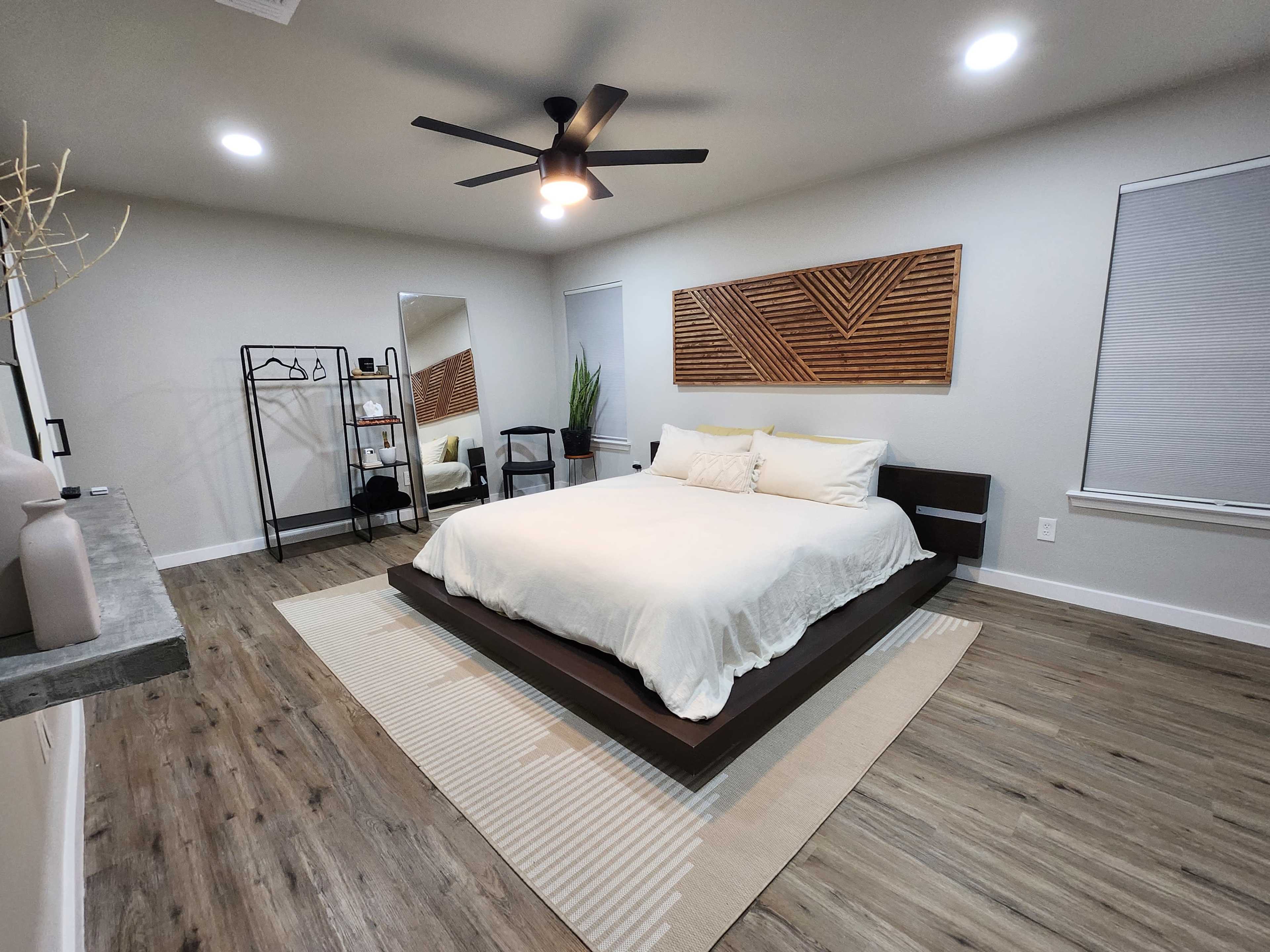 A modern bedroom featuring a large bed, a wooden accent wall, a mirror, and minimalist decor with a plant in the corner.