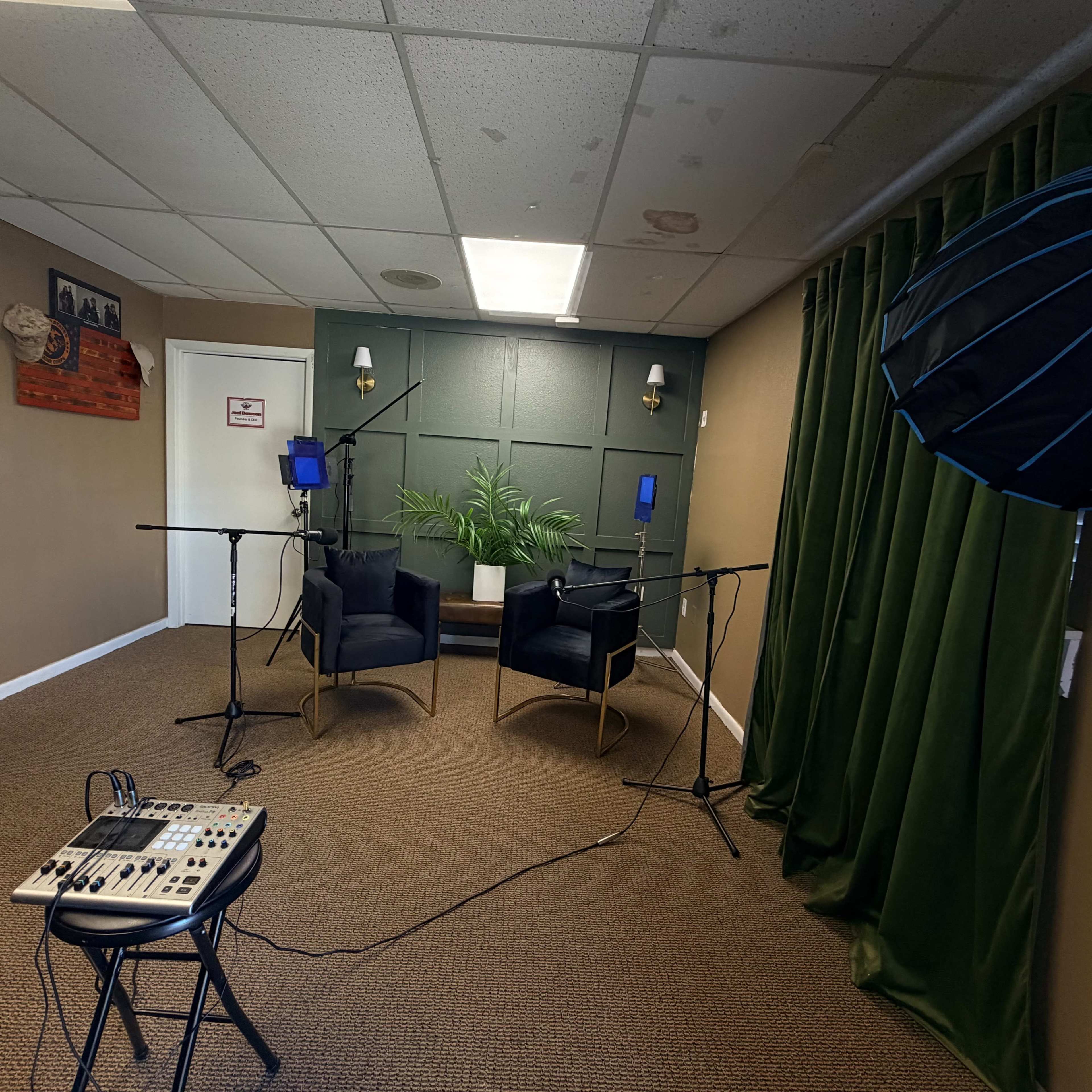 The image shows a small recording studio with two black chairs, microphones on stands, and equipment arranged around a potted plant, all set against a green curtain backdrop.
