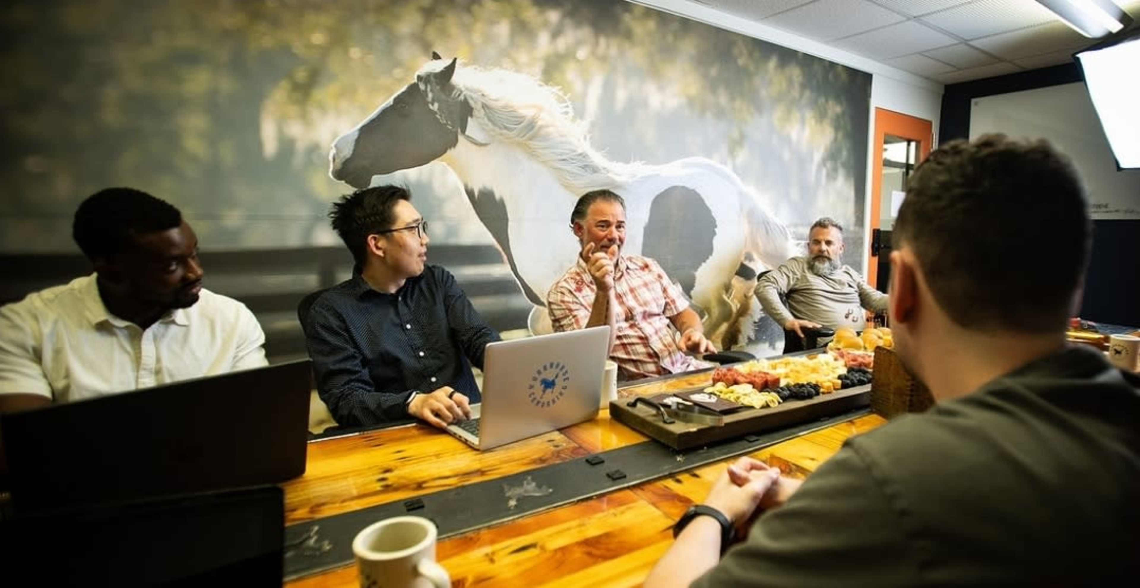 A group of five people are seated around a wooden table in a conference room, with a large mural of a horse on the wall behind them.