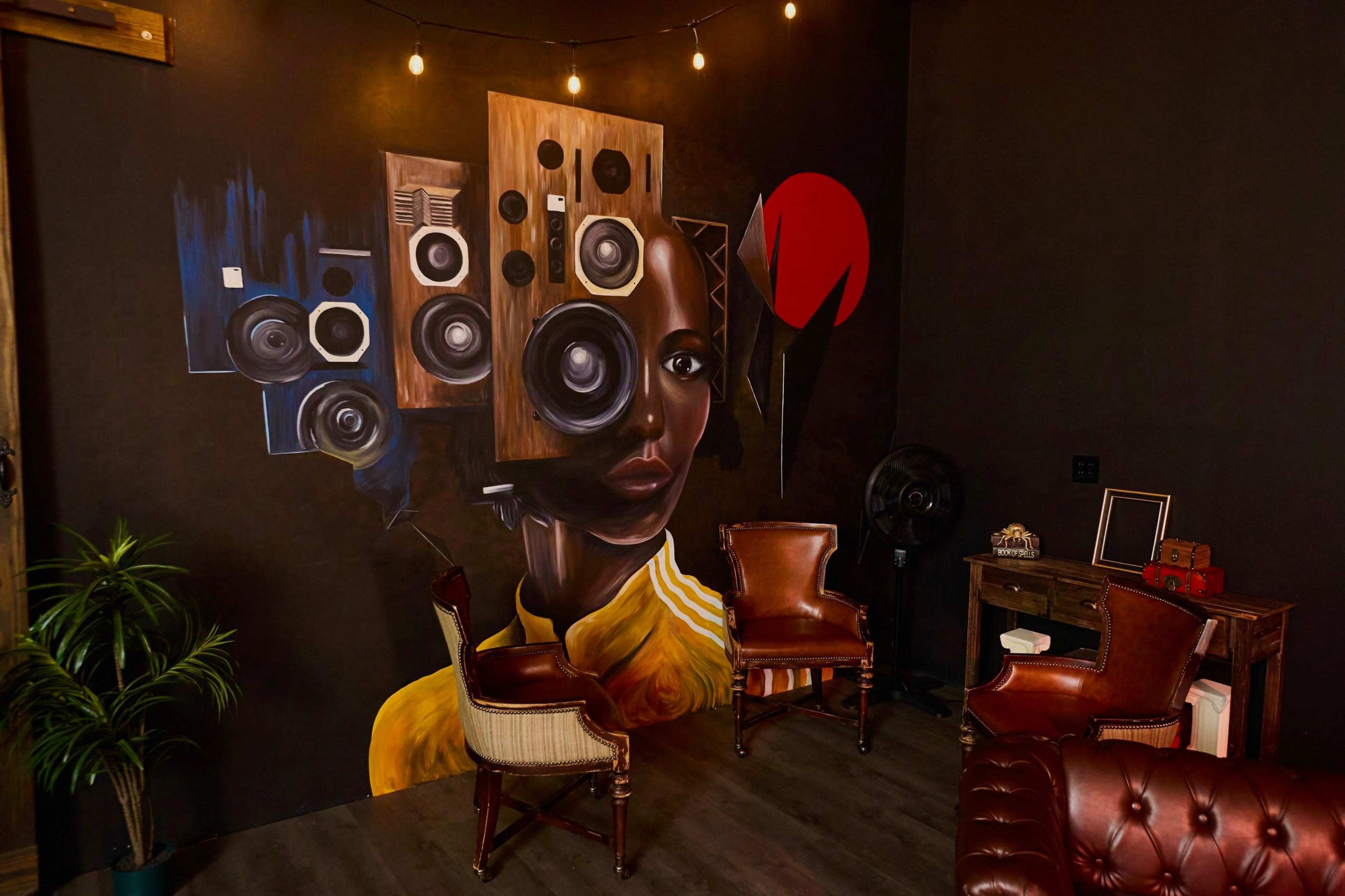 The image features a dark room with a mural of a woman alongside various speakers, two vintage chairs, and a desk in the corner.