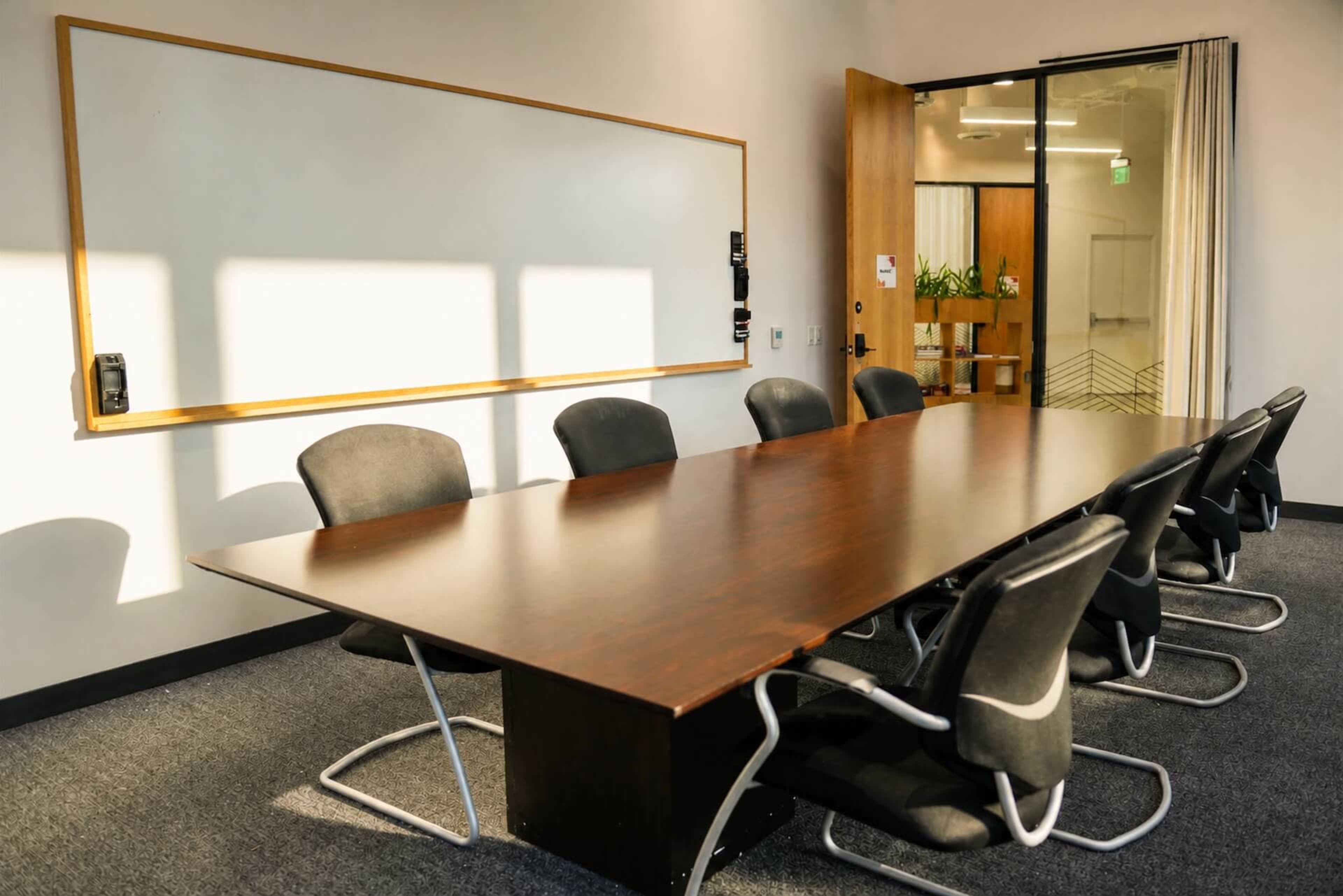 The image shows a modern conference room with a large wooden table, six black chairs, and a whiteboard mounted on the wall.