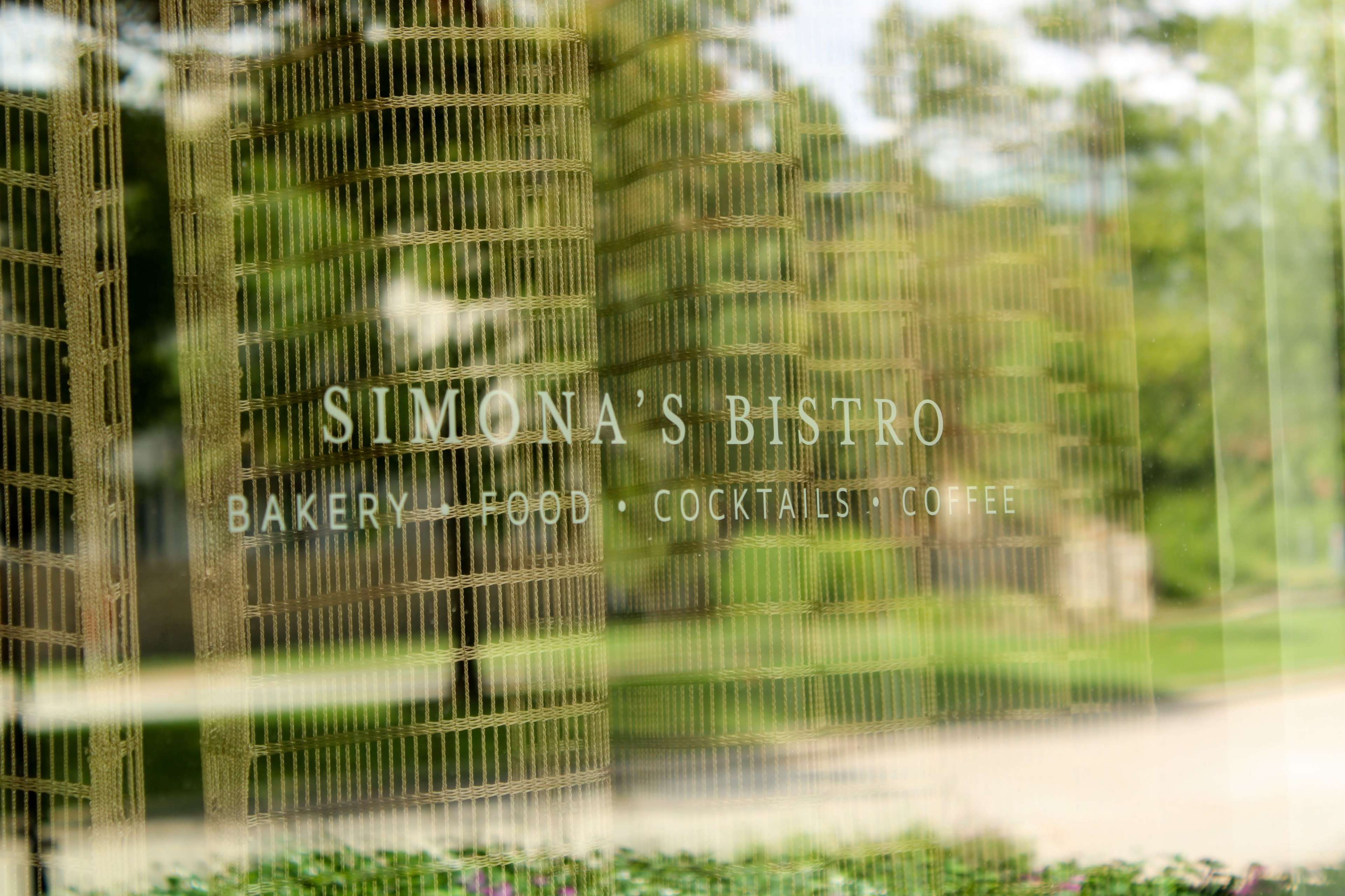 A reflective glass surface displaying the sign for "Simona's Bistro," which offers bakery items, food, cocktails, and coffee.