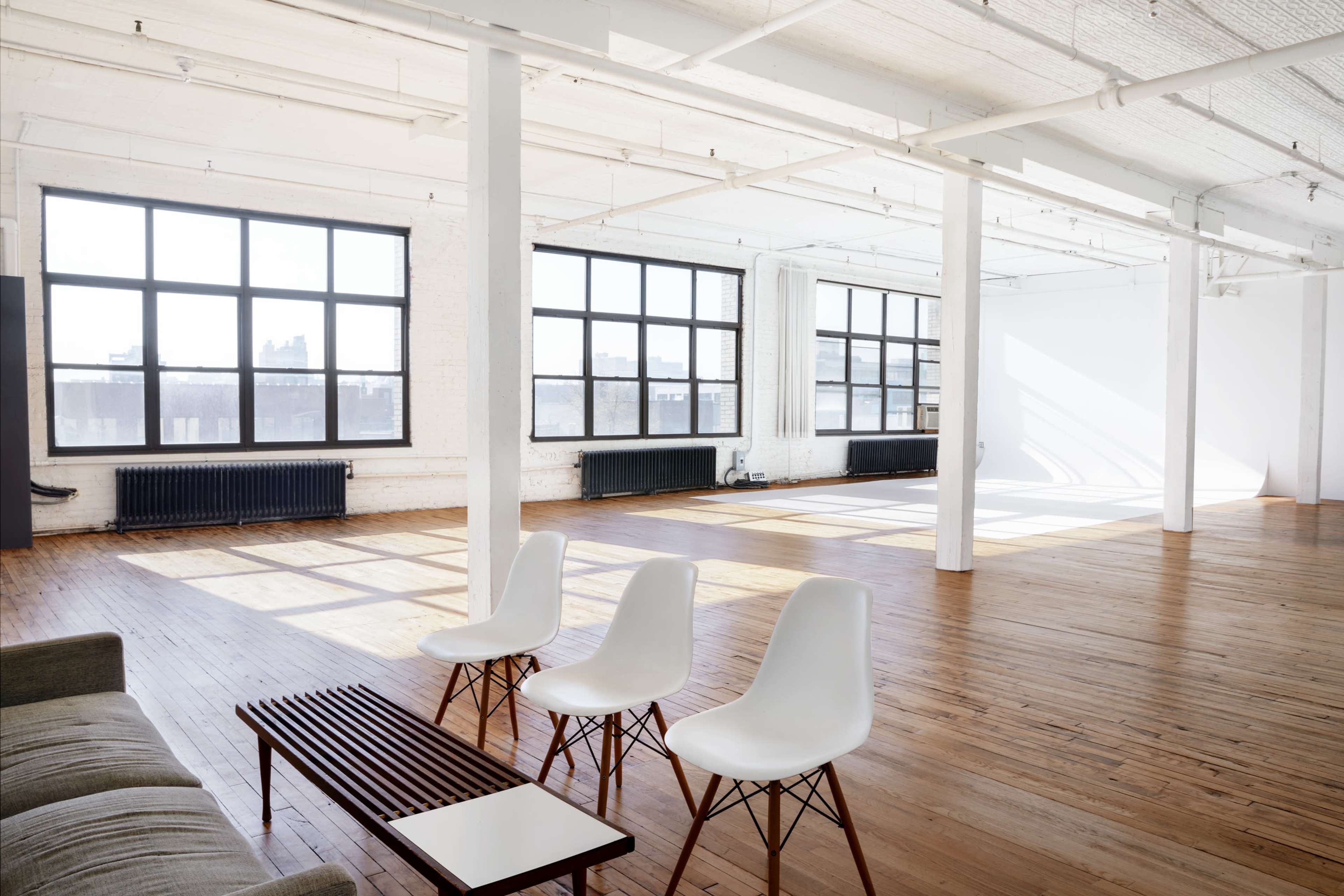 An airy, open space features large windows, wooden floors, a sofa, and three white chairs arranged around a low table.