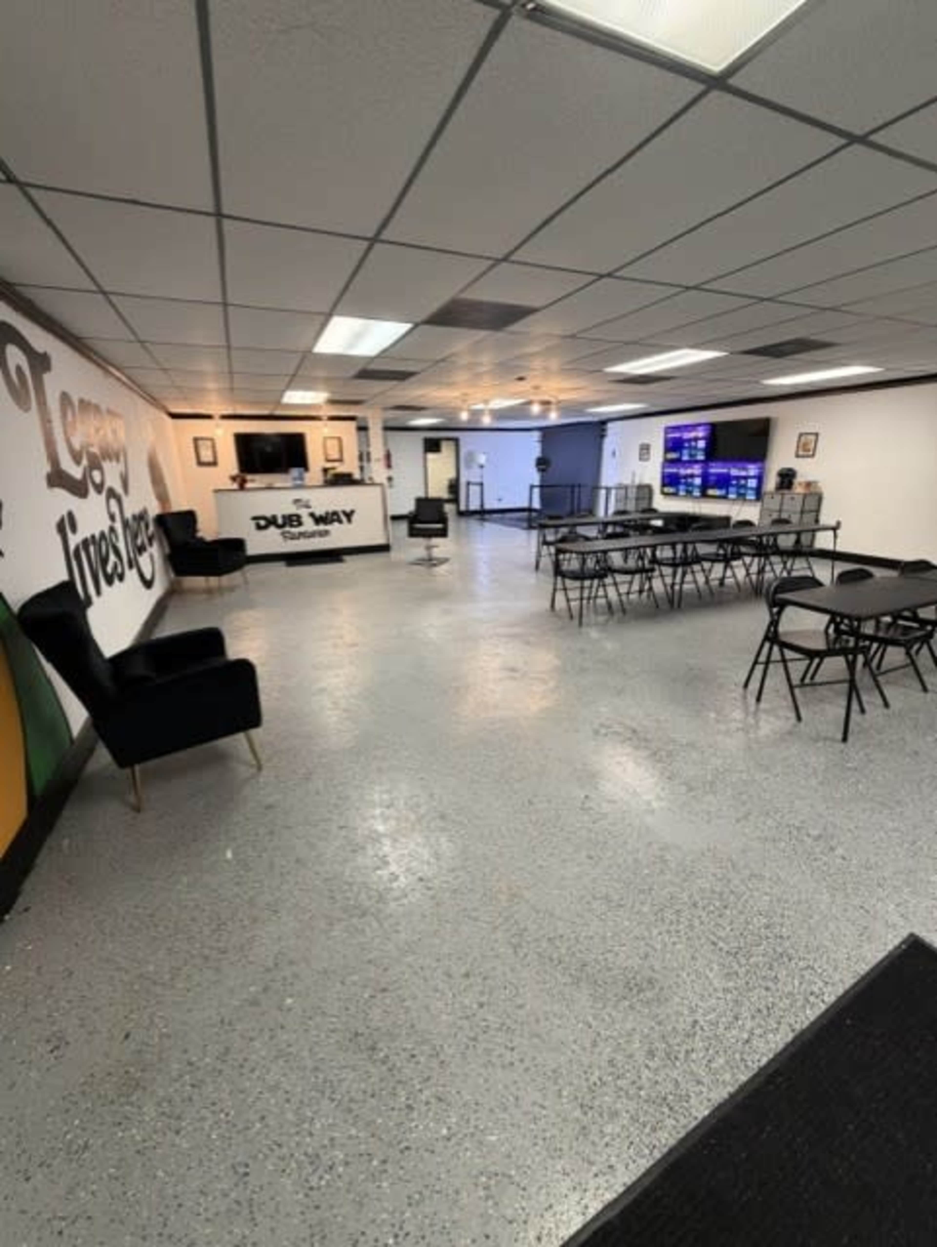 The image shows a spacious, empty commercial interior with tables and chairs set up, a service counter at one end, and large screens mounted on the walls.