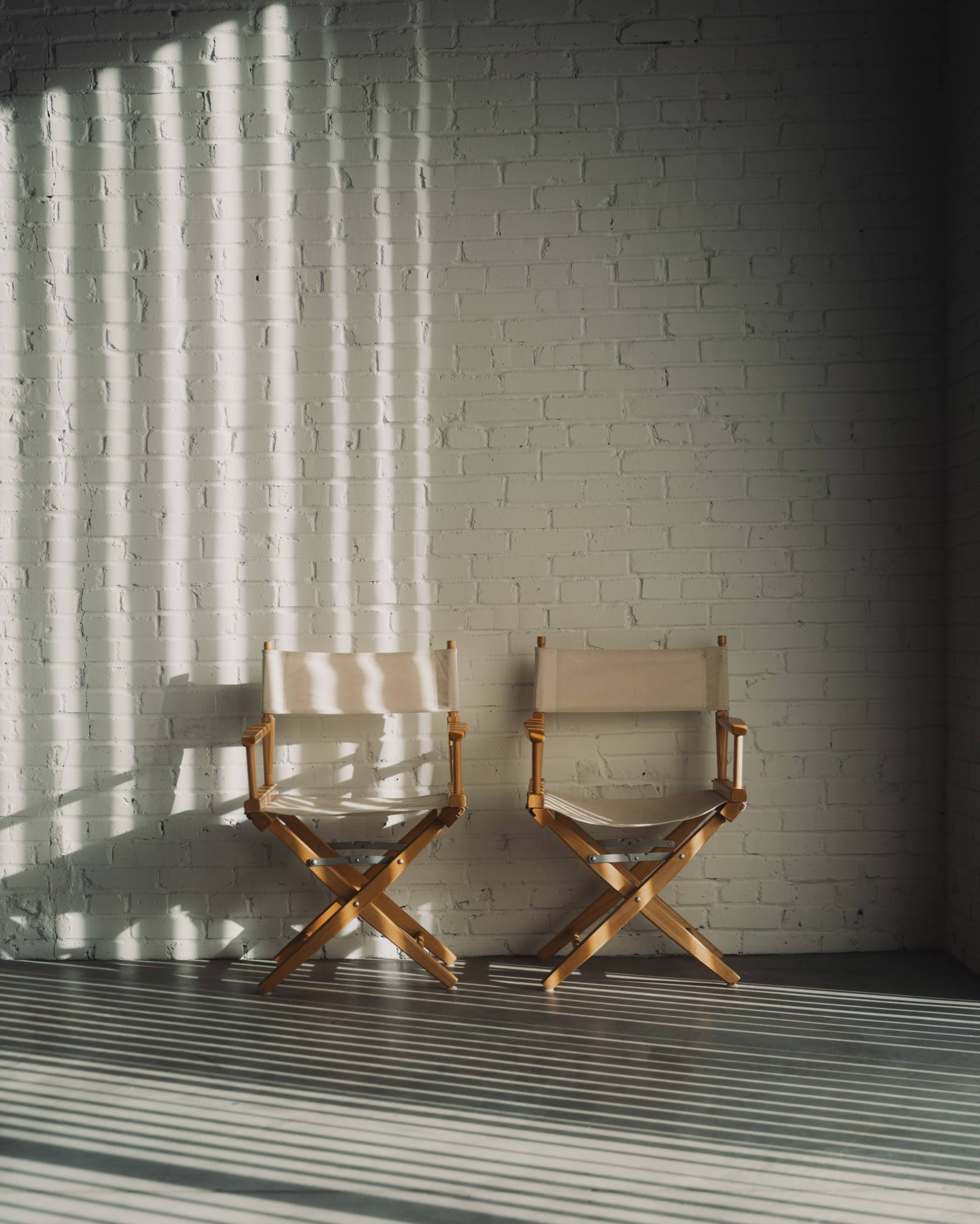 Two wooden director's chairs are positioned against a brick wall, casting shadows on the floor from the light filtering through nearby blinds.