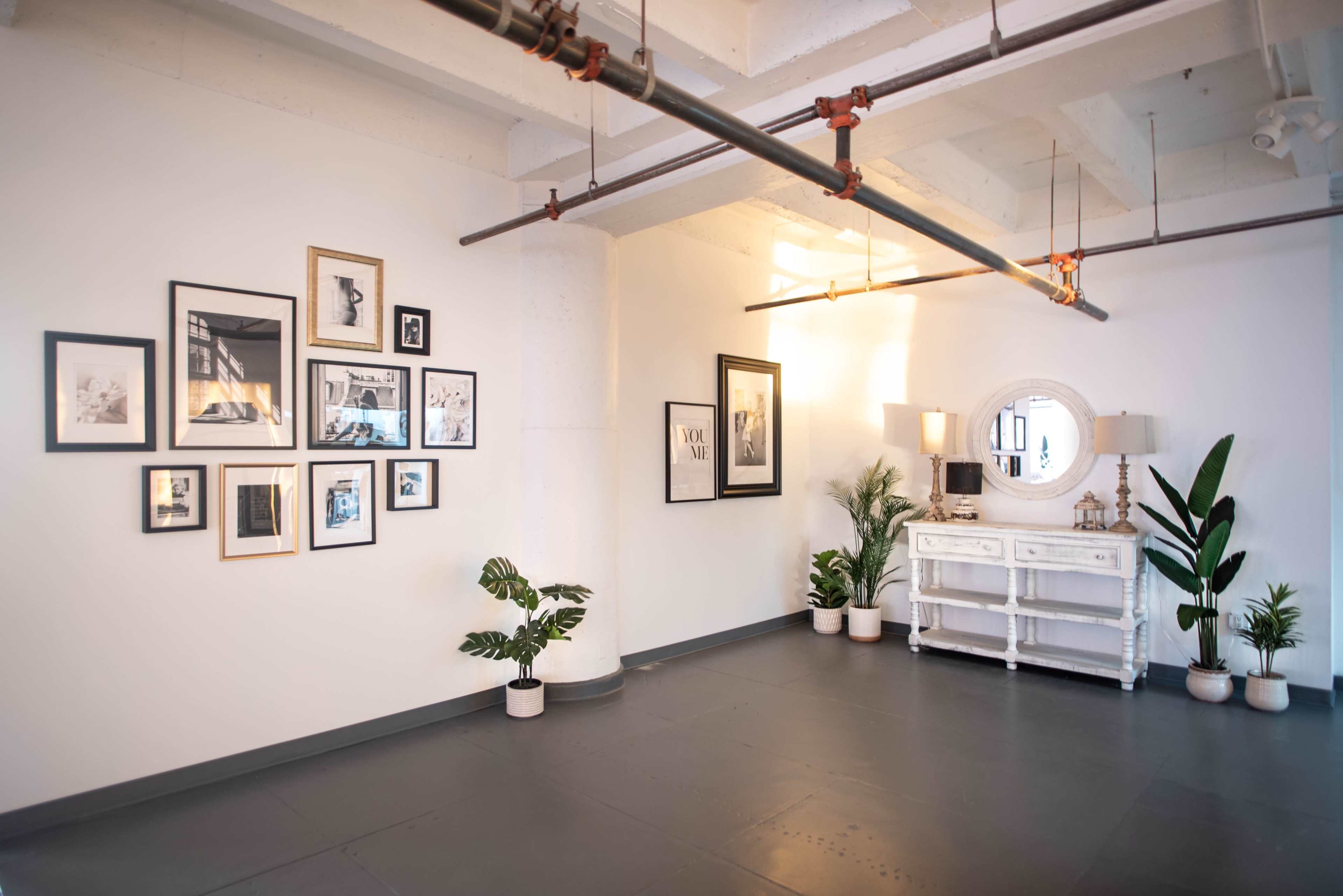 An open room with a gray floor, featuring framed photographs on the white walls, a round mirror, two lamps on a white console, and potted plants in each corner.