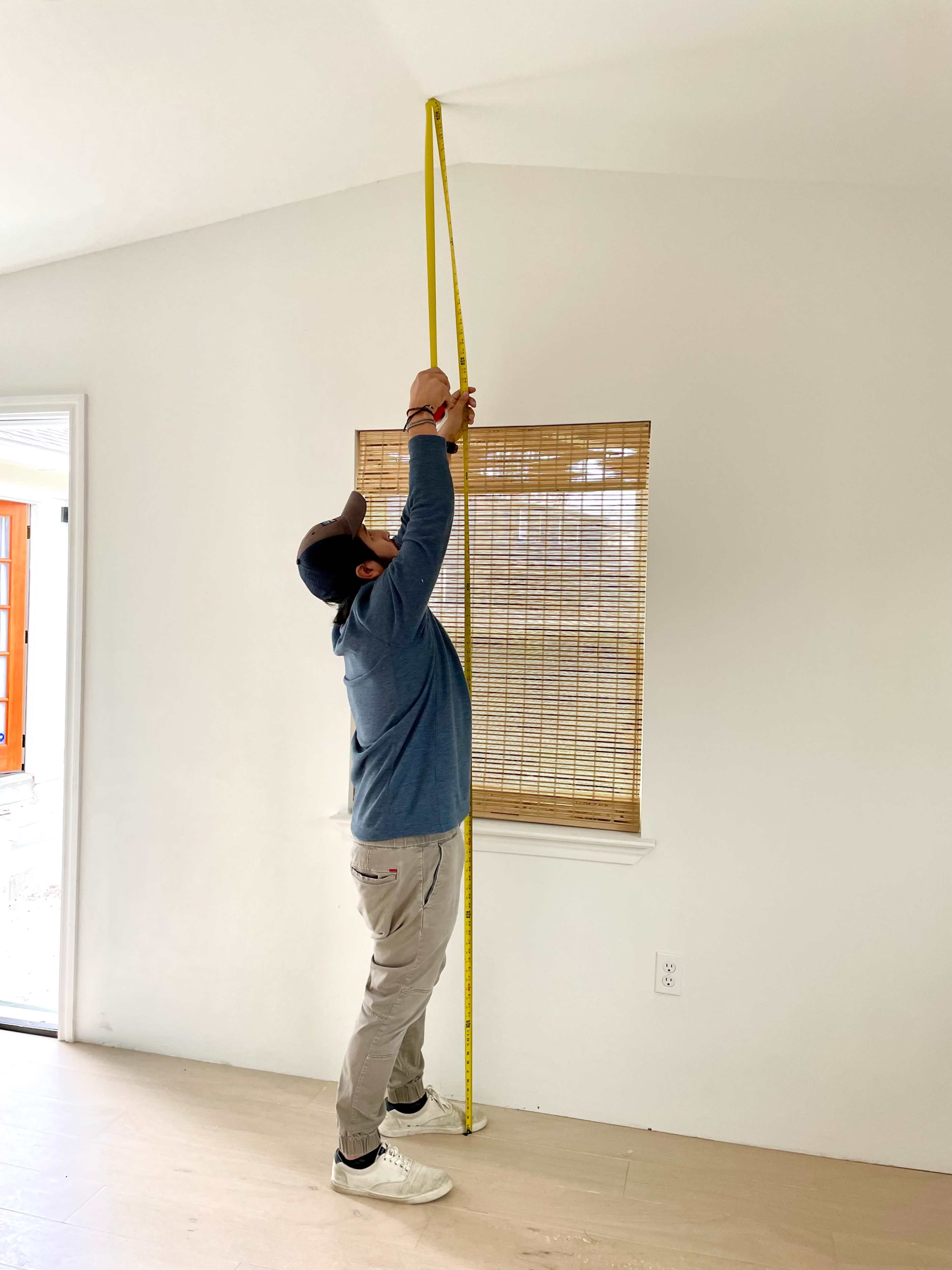 A person holds a measuring tape against the ceiling in a room with a window covered by a bamboo blind.