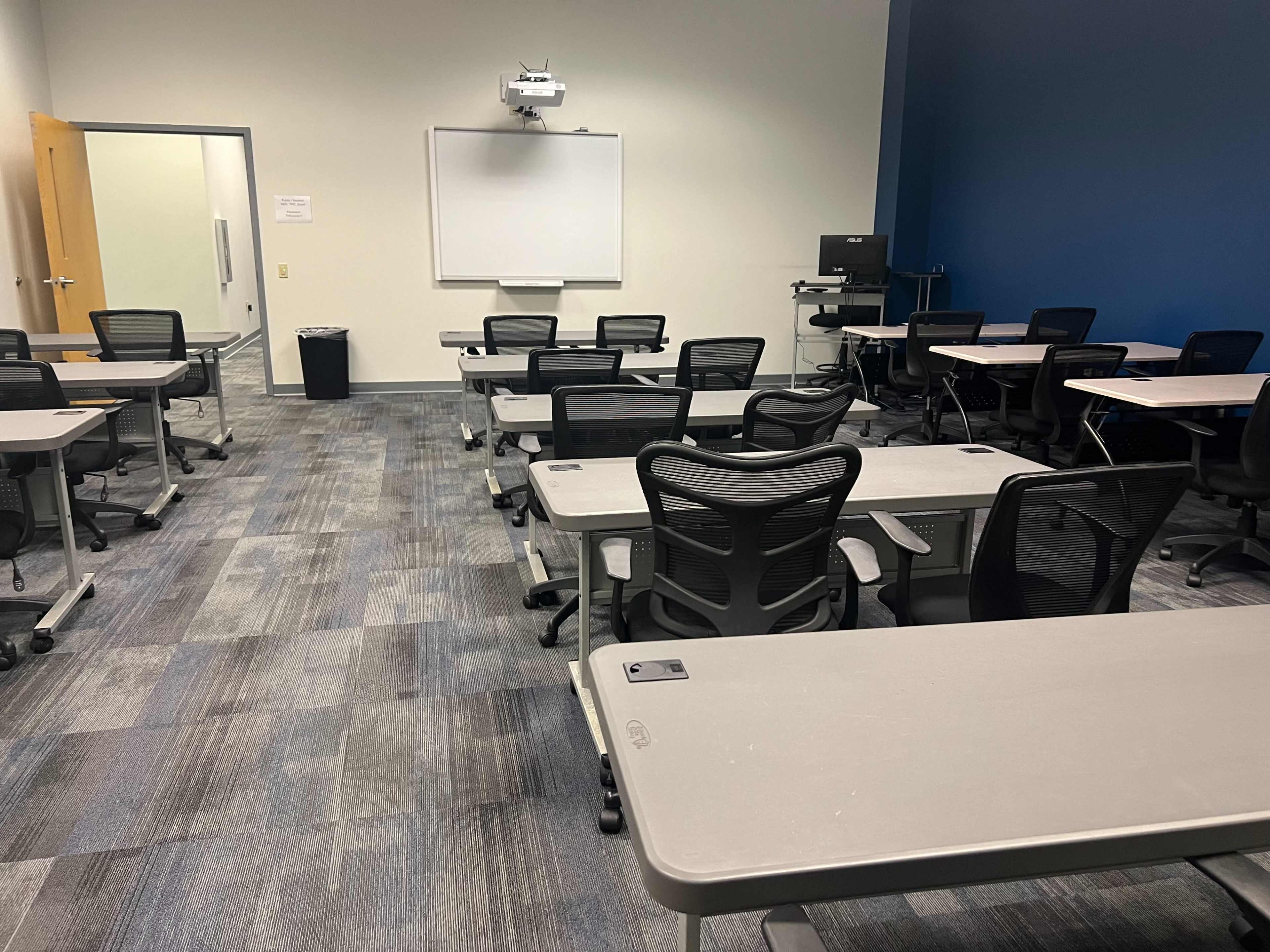 A classroom is arranged with multiple movable desks and chairs facing a whiteboard and projector.