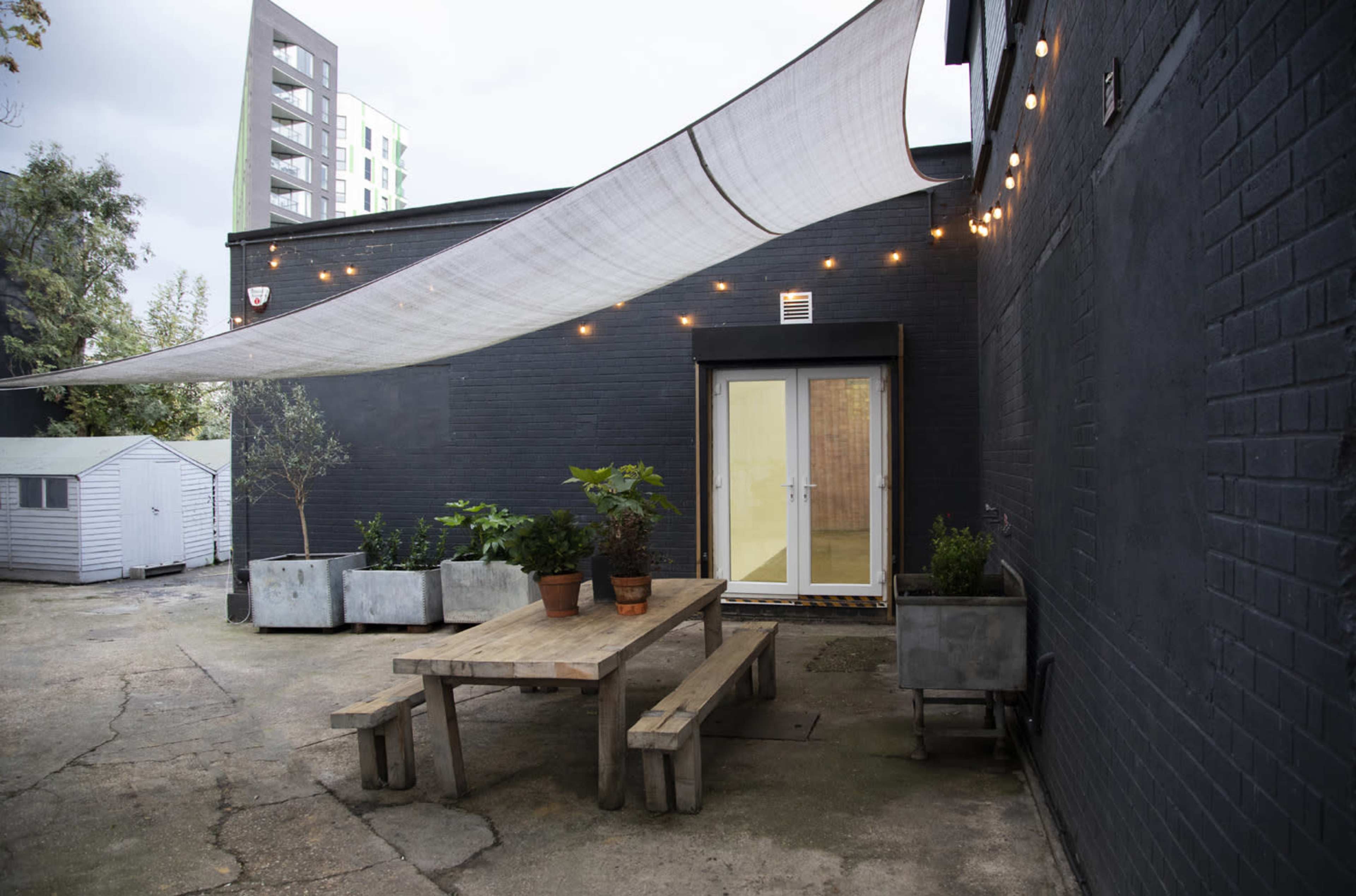 A wooden table with benches is positioned under a large fabric canopy and string lights, next to a black wall and several potted plants.