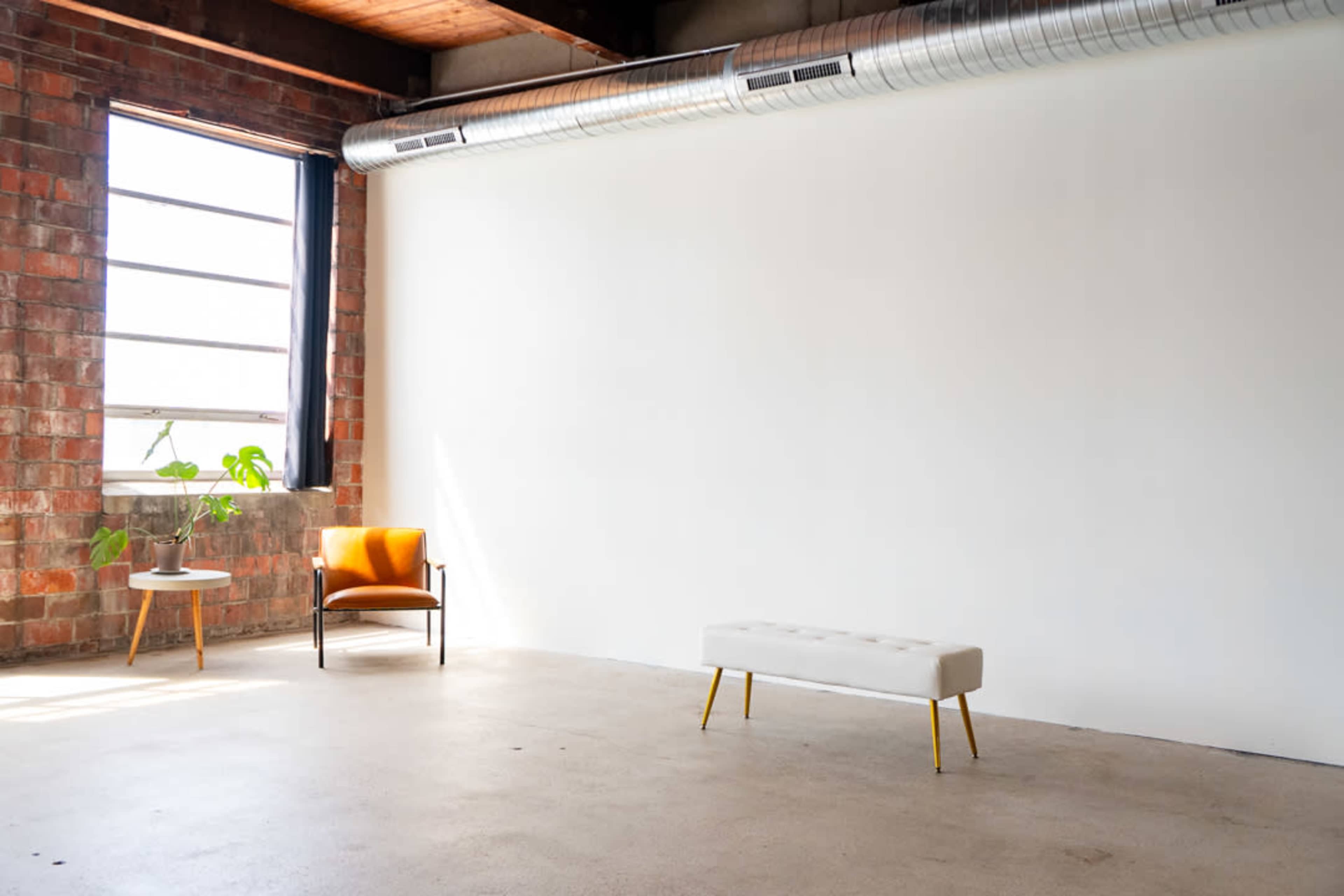 A bright, minimalist room features a large window, a potted plant, an orange armchair, and a white bench against a plain white wall.