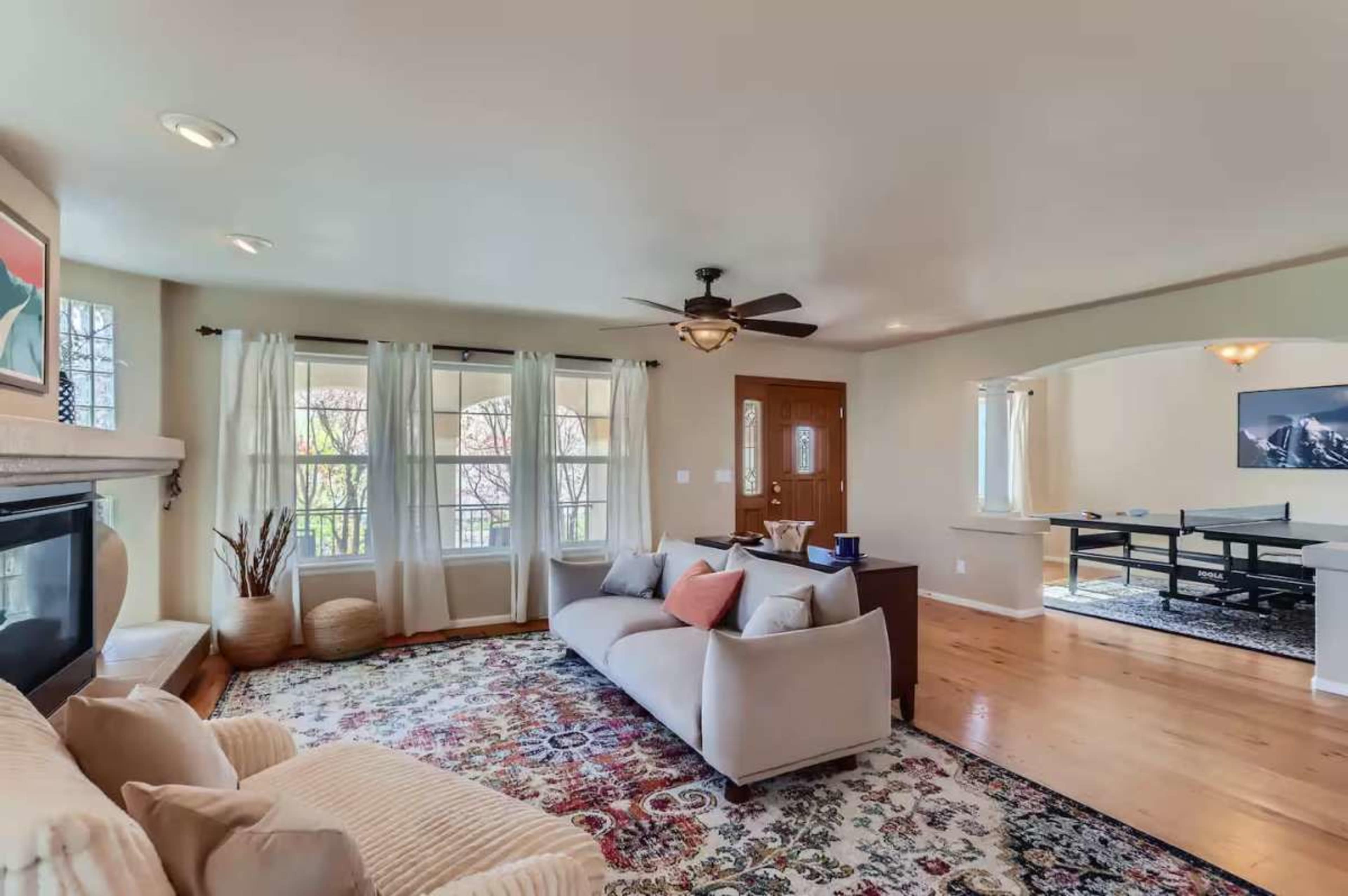 The image shows a living room with a light-colored sofa, a patterned rug, and large windows letting in natural light.