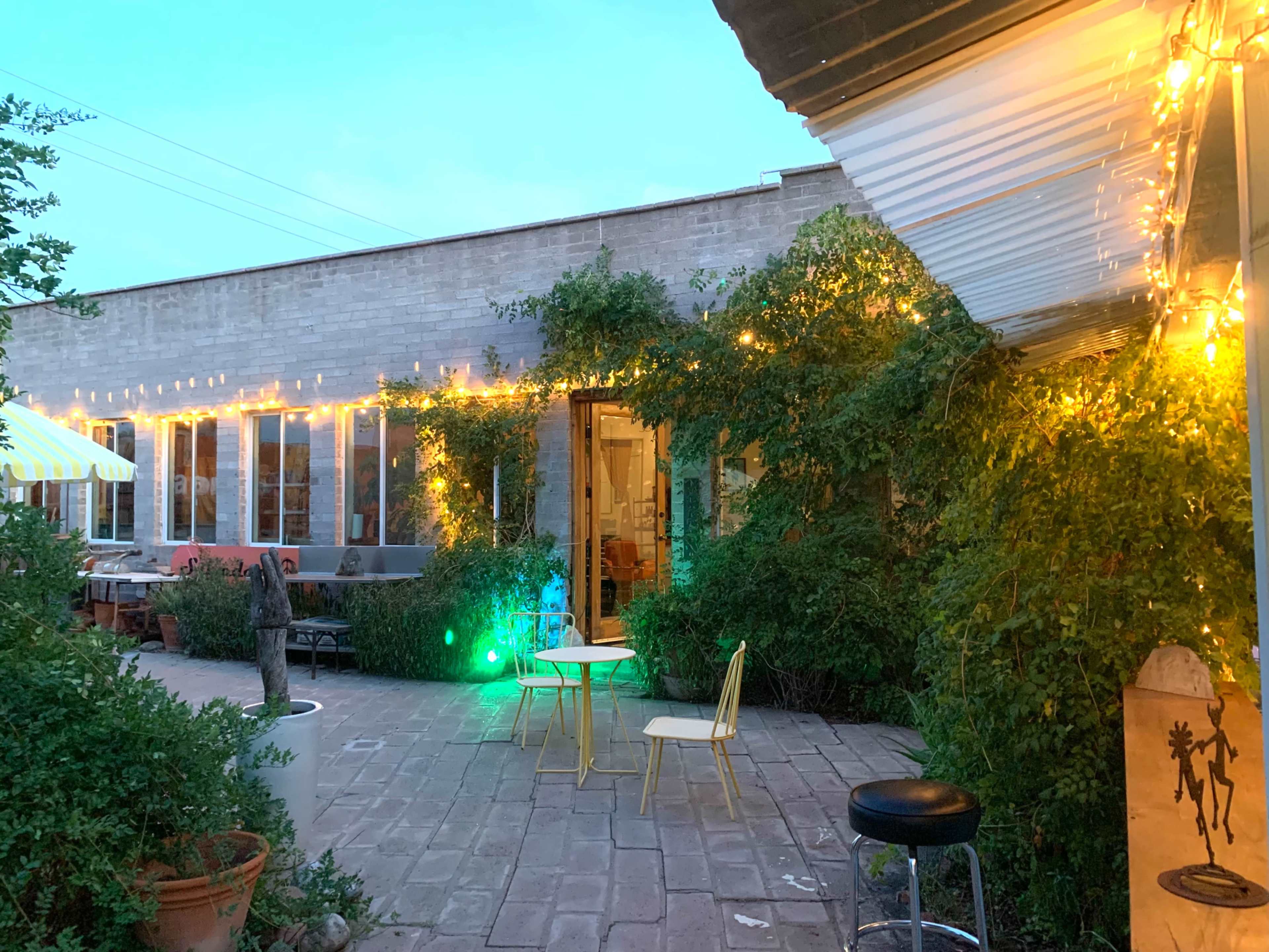 The image shows an outdoor seating area with string lights, green foliage, and two chairs arranged around a small table in front of a building.