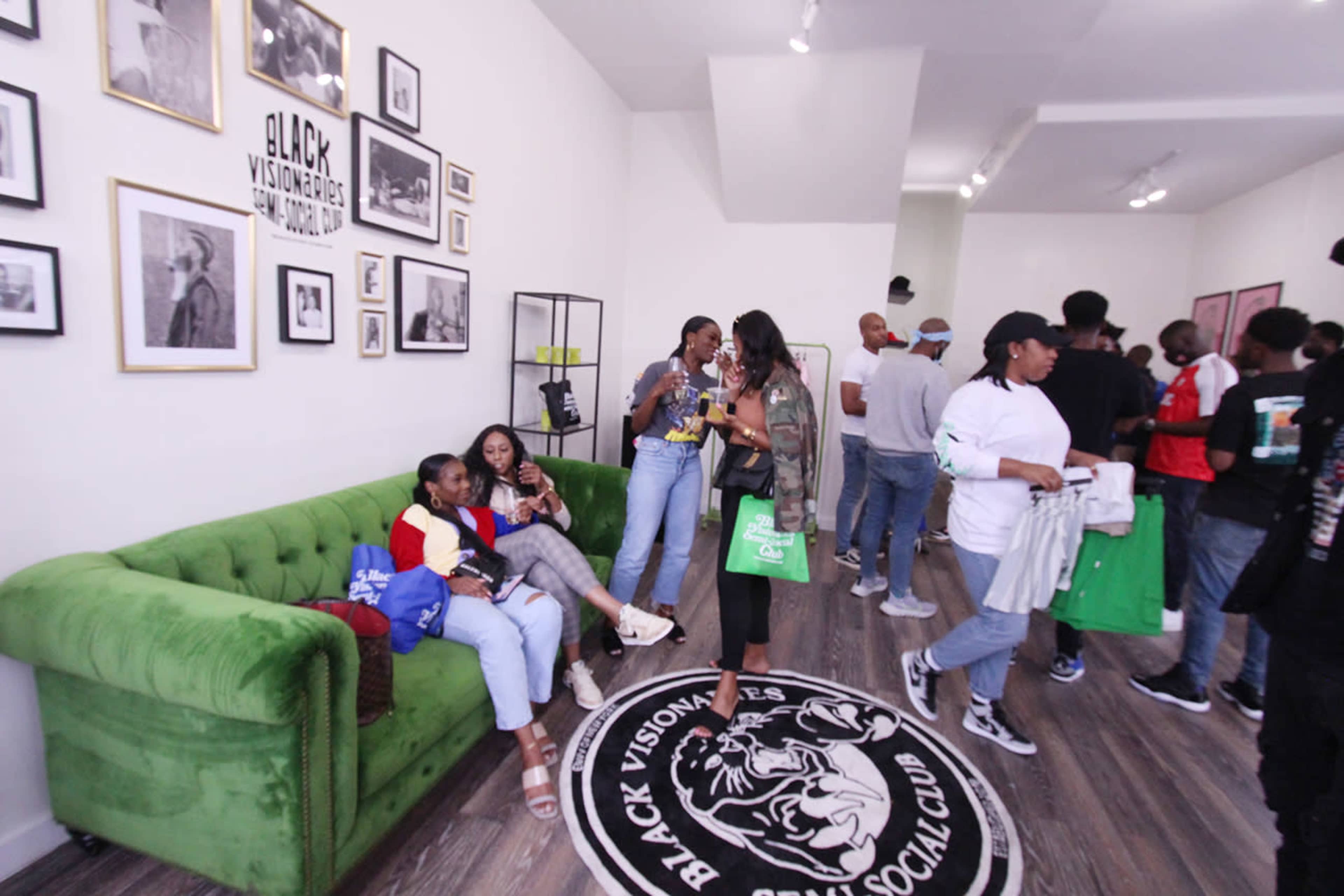 A group of people socializes in a brightly lit room adorned with framed photographs and a green sofa.