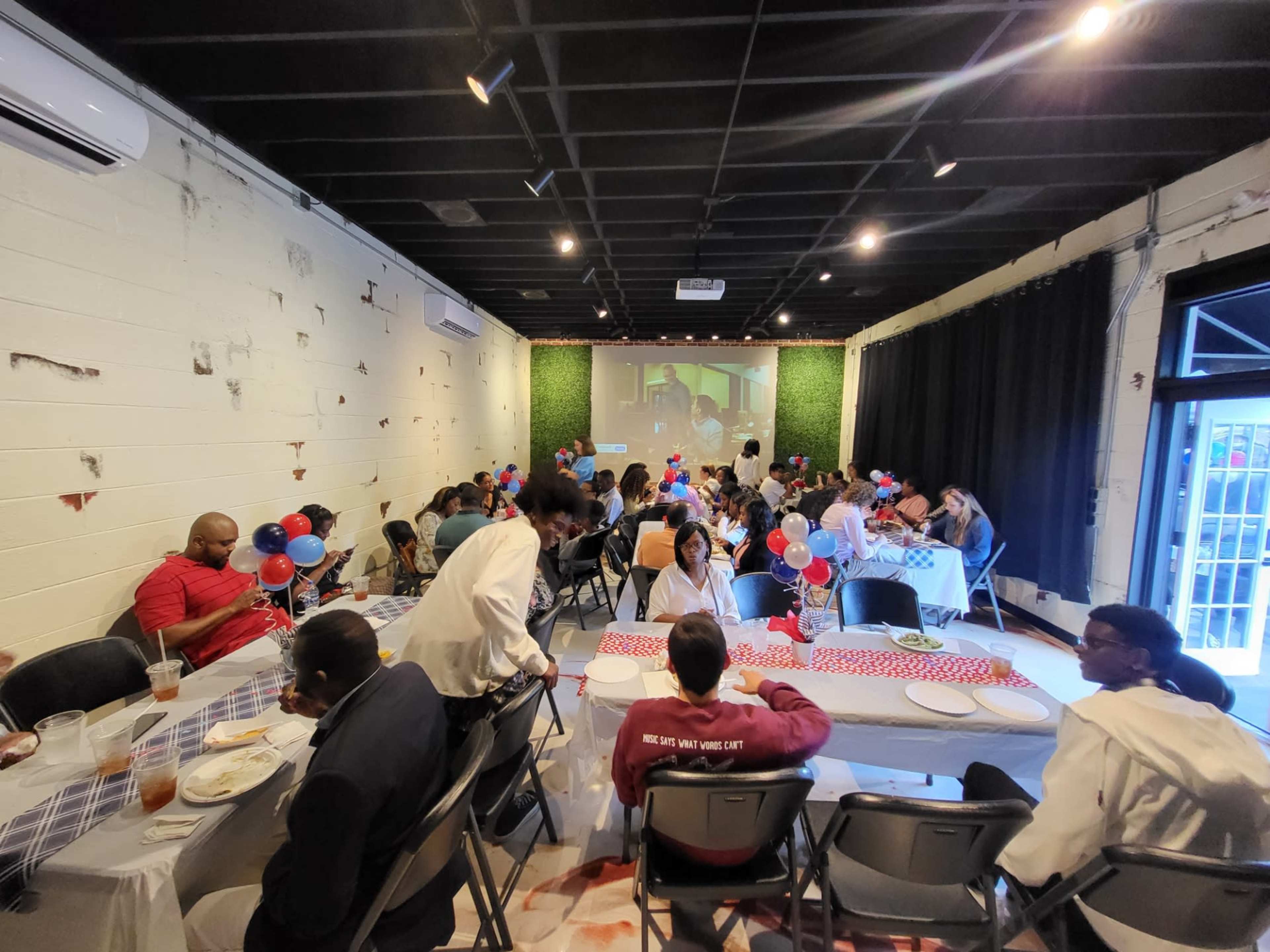 A lively gathering in a decorated room with tables set for a group celebration, featuring balloons and a large screen displaying a presentation.
