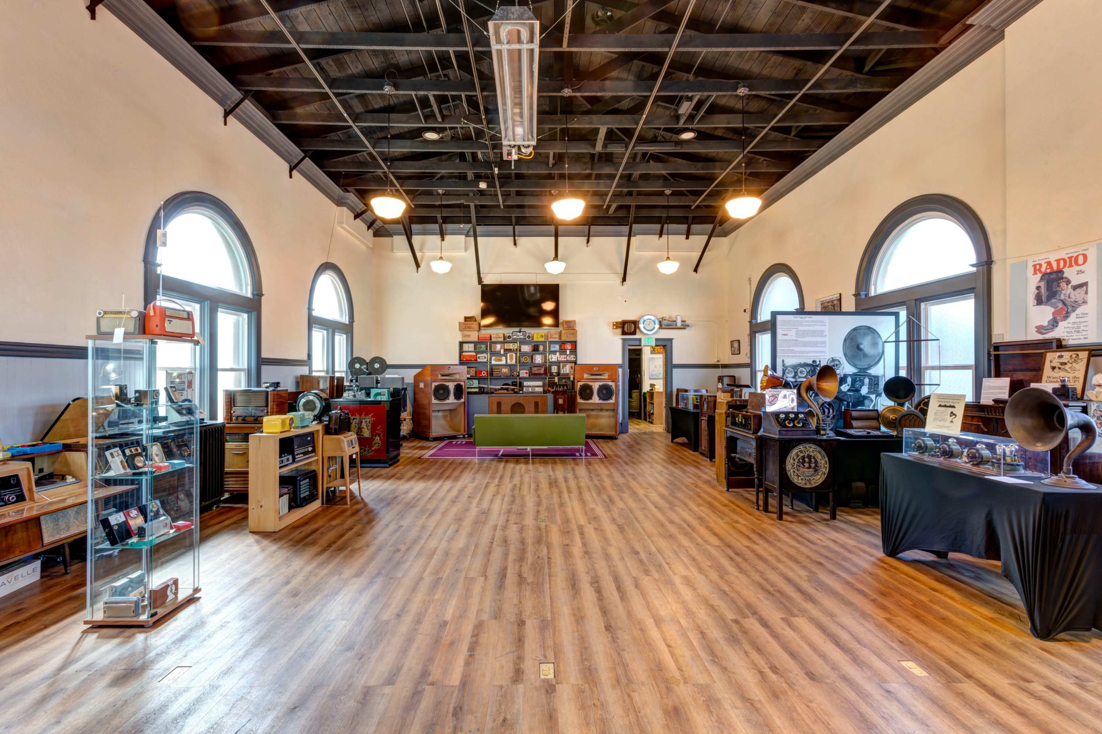 The image shows a spacious museum-like room with vintage radios and phonographs displayed on wooden shelves and tables, featuring large windows and a wooden floor.