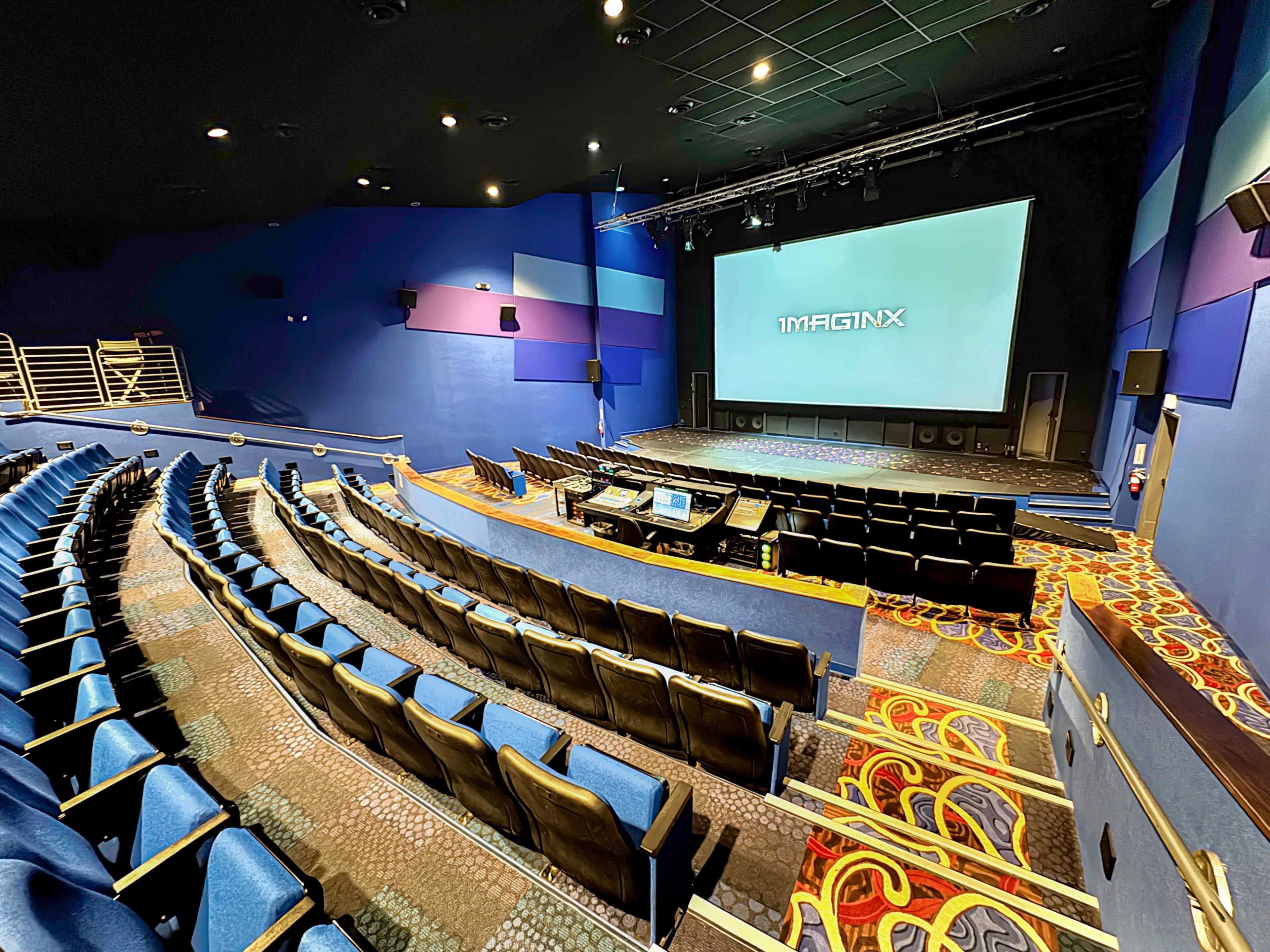 The image shows a modern cinema auditorium with tiered seating, a large screen at the front, and a control area for audio-visual equipment.