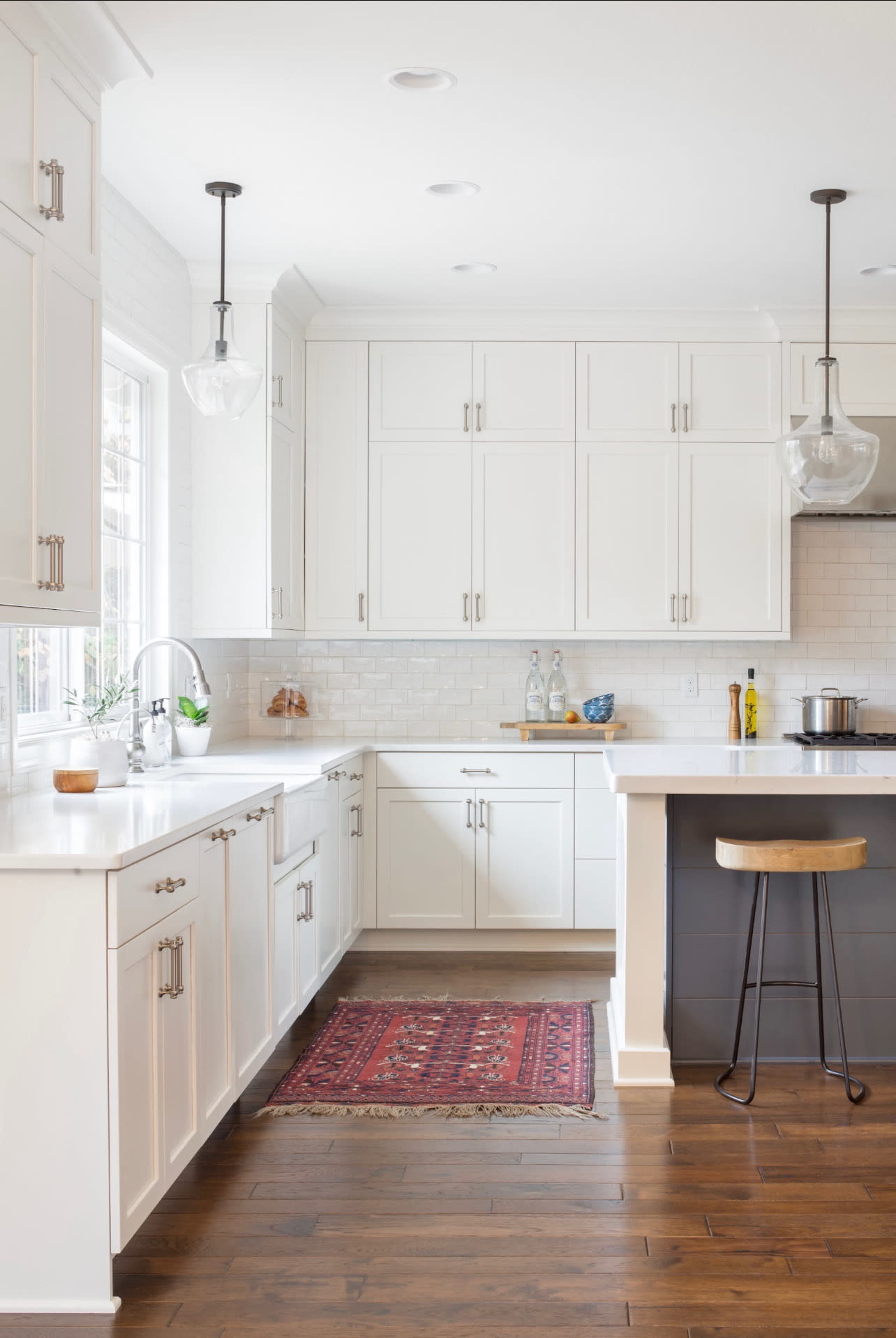A bright kitchen features white cabinets, a marble countertop, and wooden flooring, with a decorative rug and modern bar stool.