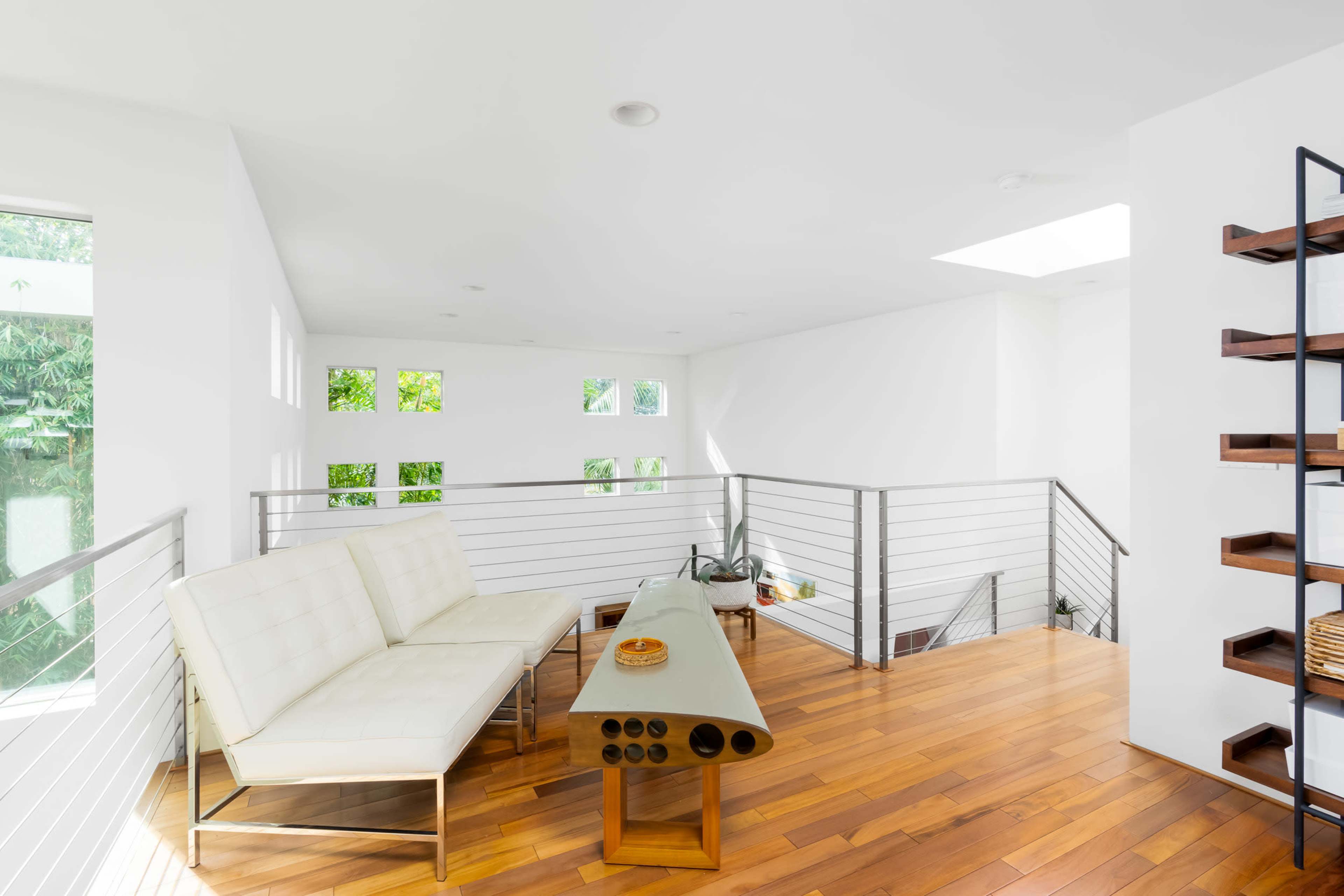 A bright, modern loft space features a white sofa and a wooden coffee table, surrounded by large windows and sleek metal railings.