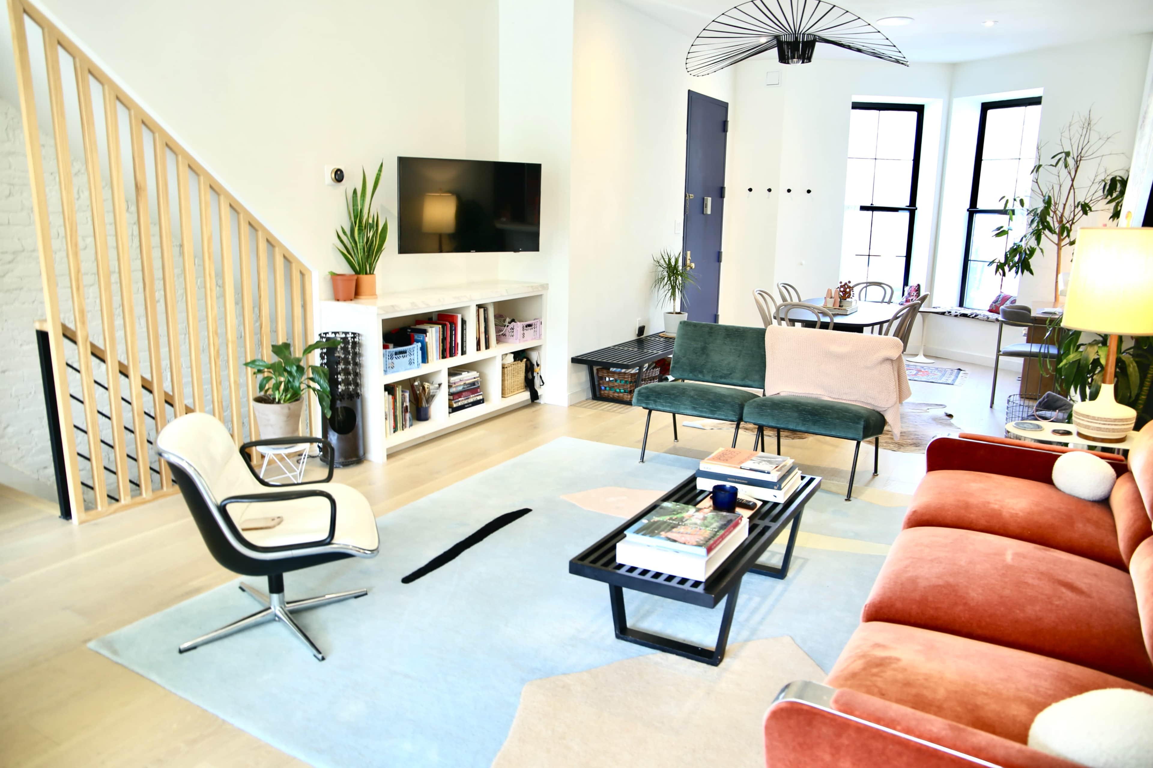 The image shows a bright living room featuring a modern design with a mix of seating options, a television mounted on the wall, and decorative plants.