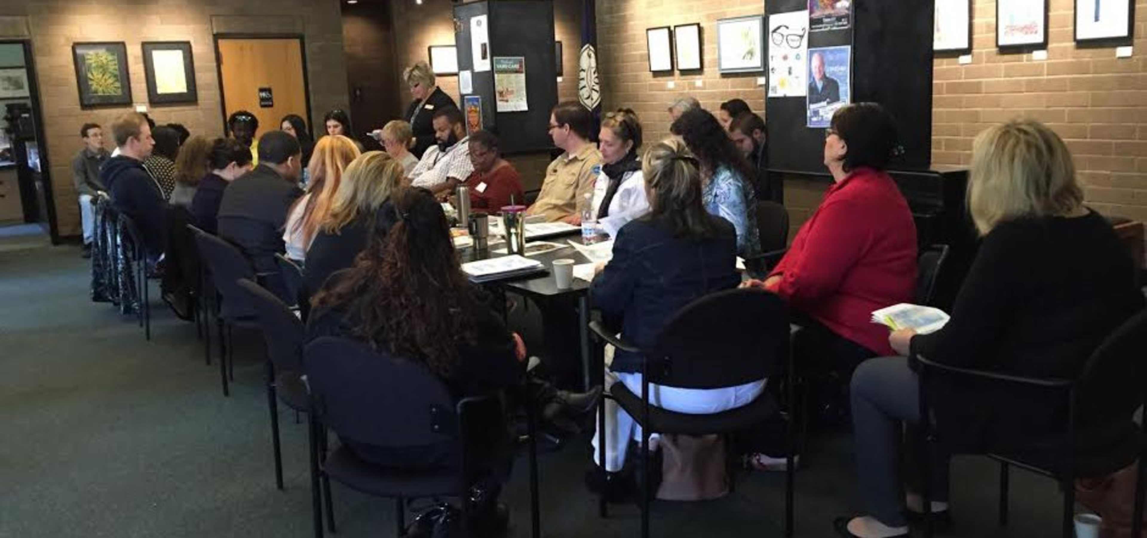 A group of people sits around a long table in a meeting room, with a person standing at the head of the table speaking to the attendees.