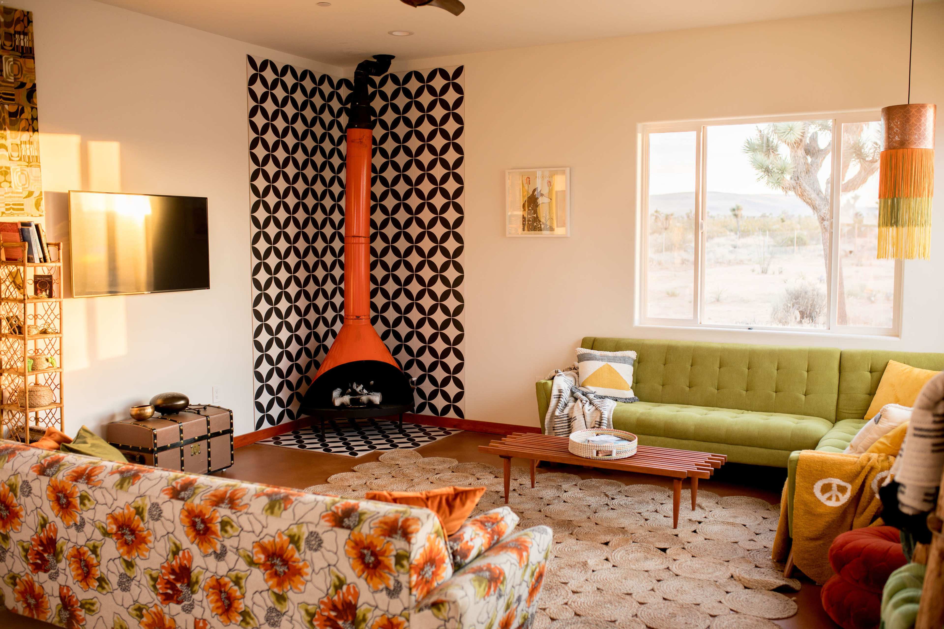 A modern living room with colorful furniture, including a green sofa and a floral-patterned couch, centered around a black and white patterned rug and an orange chimney fireplace.
