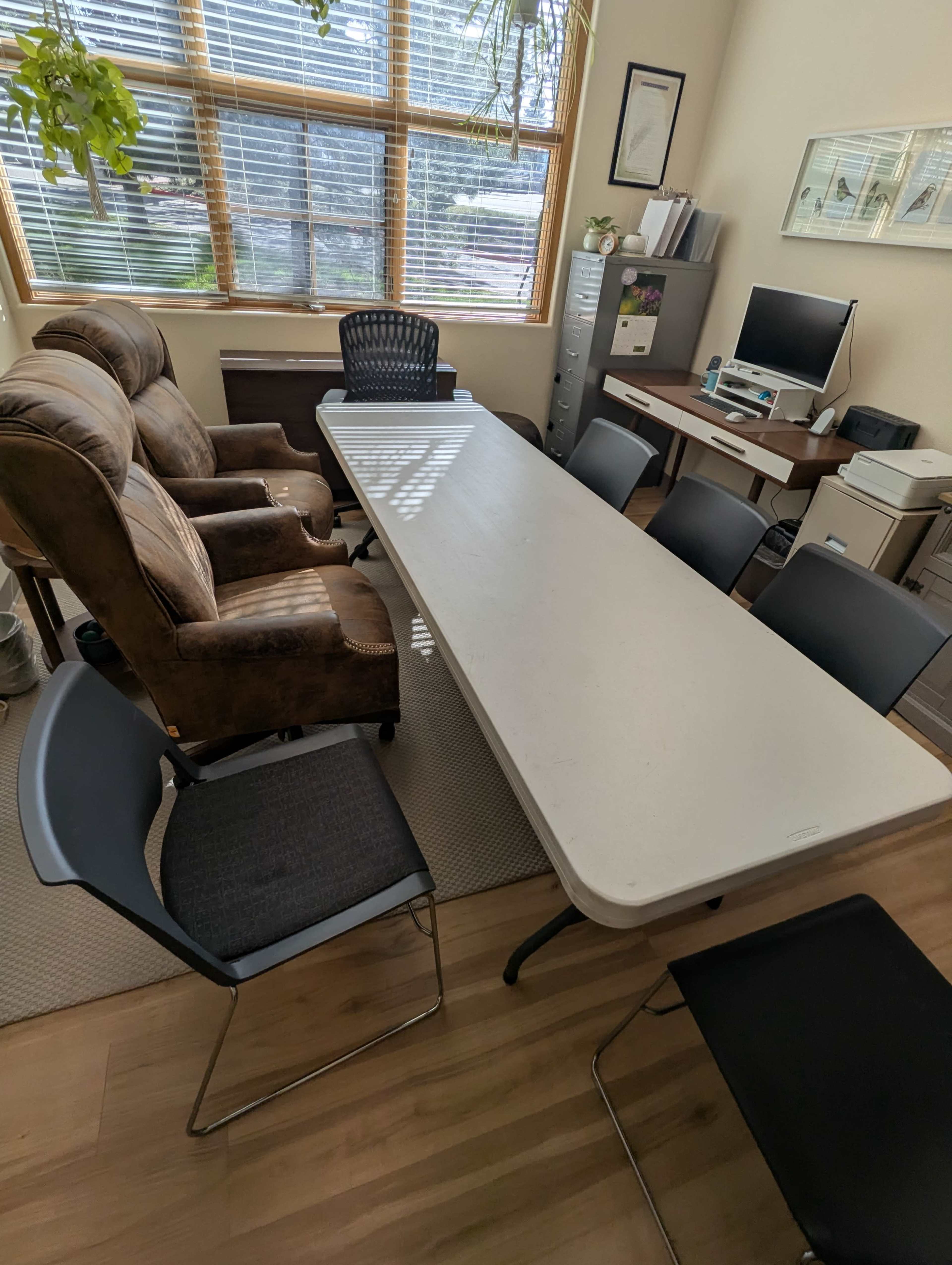 The image shows an office space with a long table surrounded by several chairs, two armchairs in the corner, and a desk with a computer and filing cabinet.