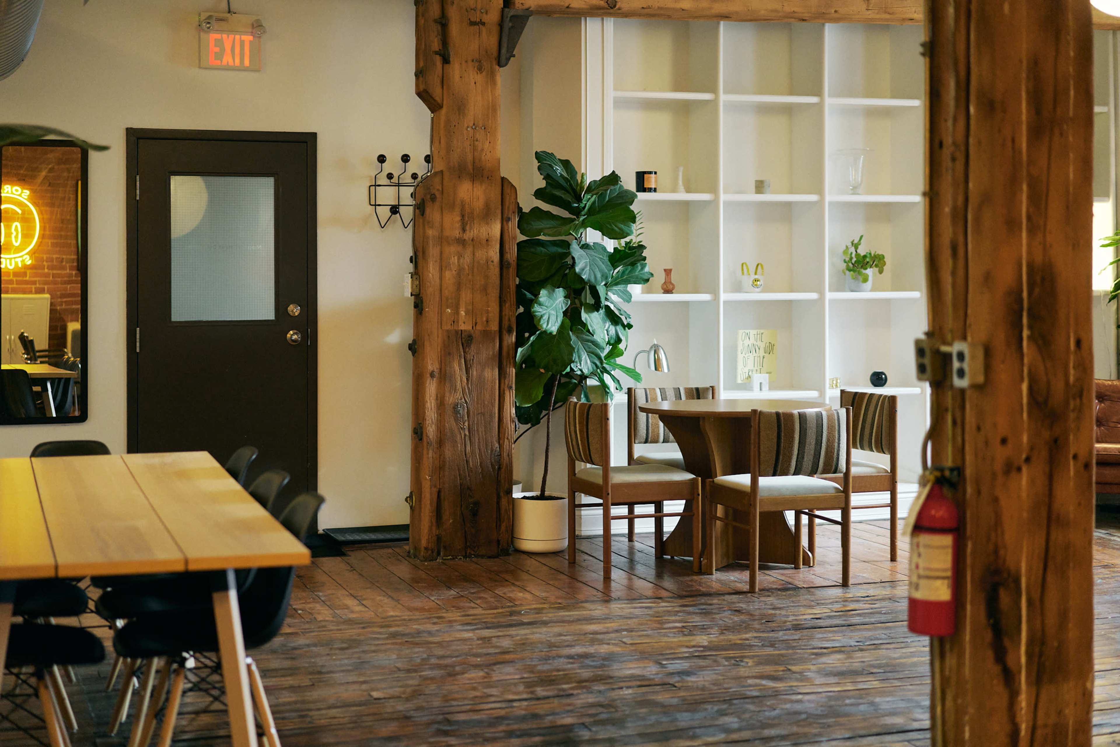 The image shows a modern interior space featuring wooden beams, a dining table with chairs, a bookshelf, and potted plants, along with an exit sign and fire extinguisher on the wall.