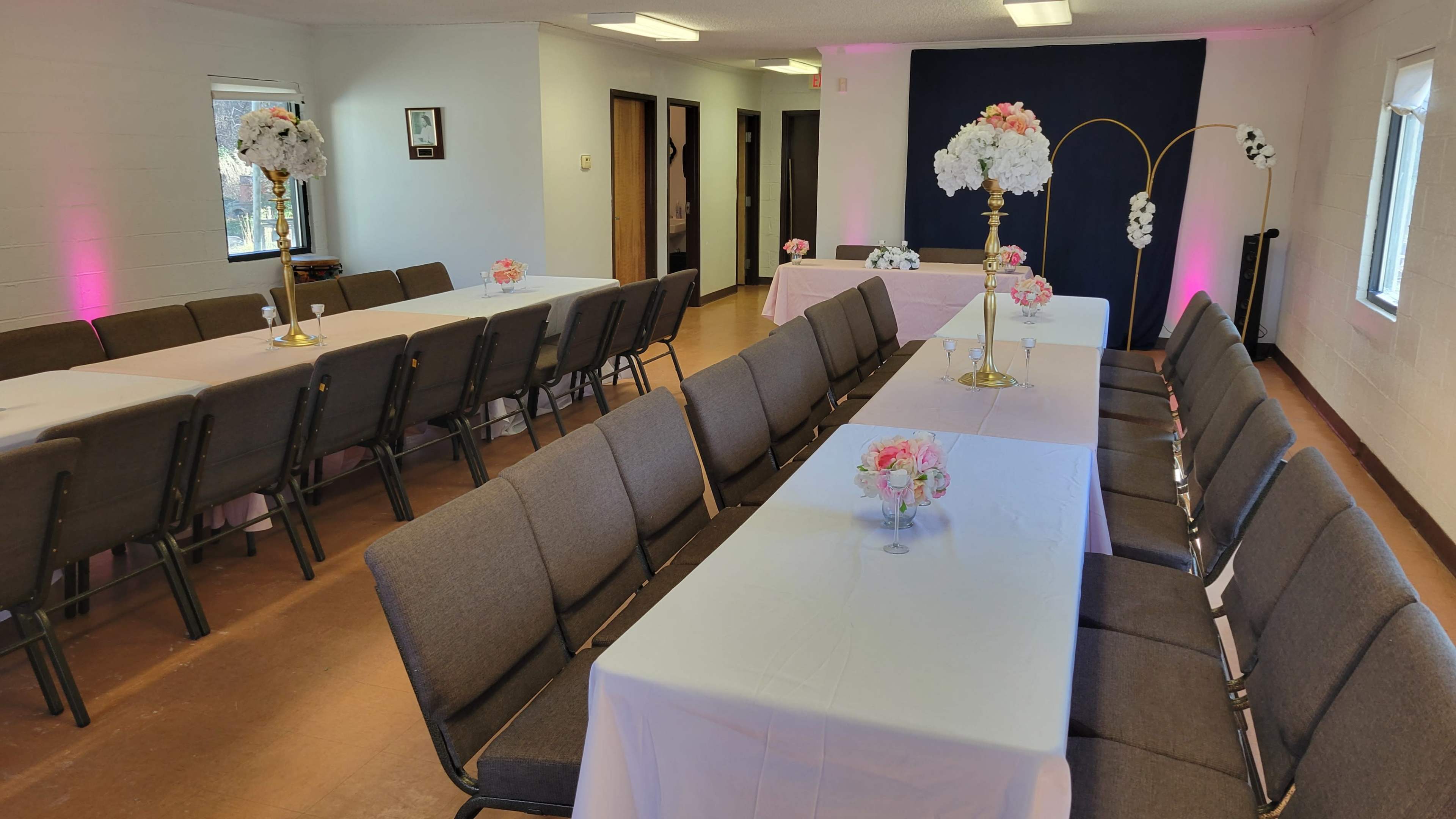 The image shows a well-organized banquet hall with neatly arranged tables covered in white tablecloths, decorated with floral centerpieces, and framed by a dark blue backdrop.