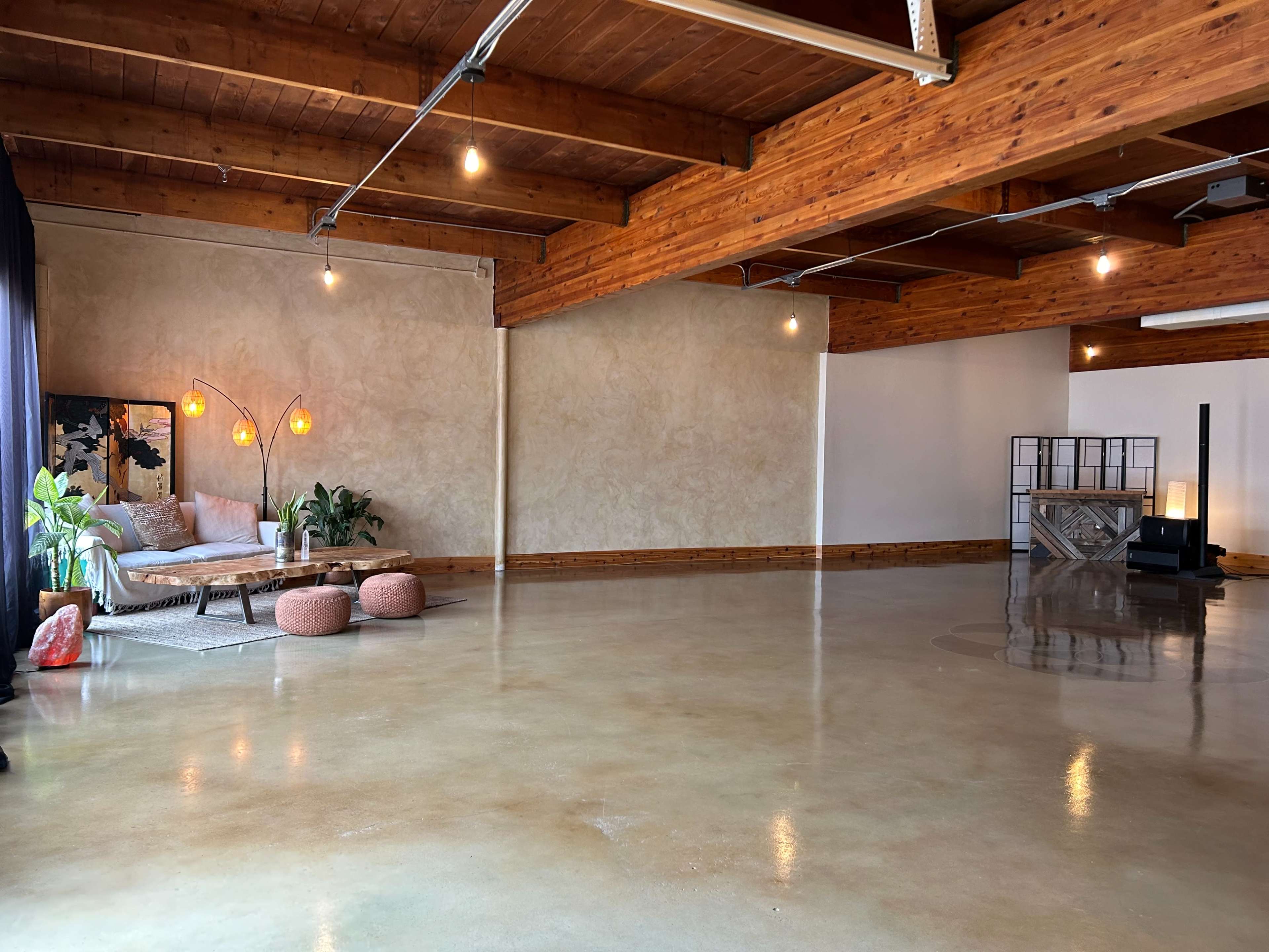 A spacious, minimalist studio with wooden beams, polished concrete flooring, and a cozy seating area featuring a light-colored couch, floor lamps, and decorative plants.