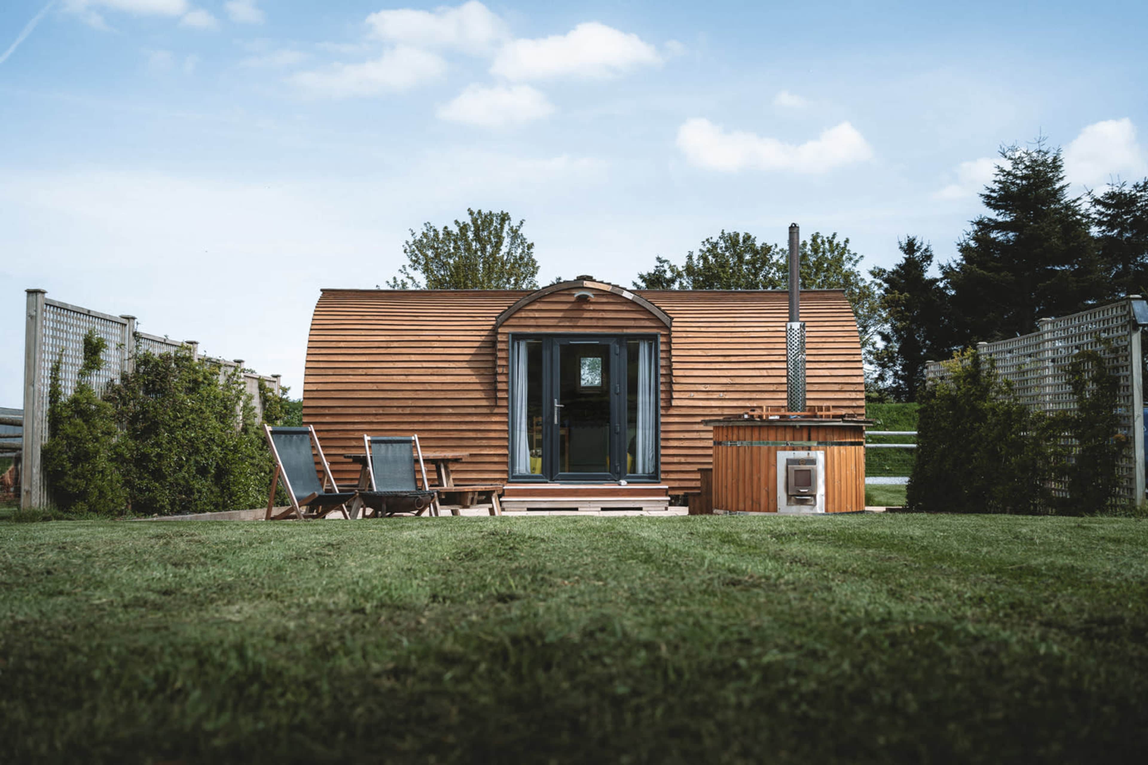 A wooden cabin with a curved roof is situated on a grassy area, featuring two lounge chairs in front and a small hot tub on the side.