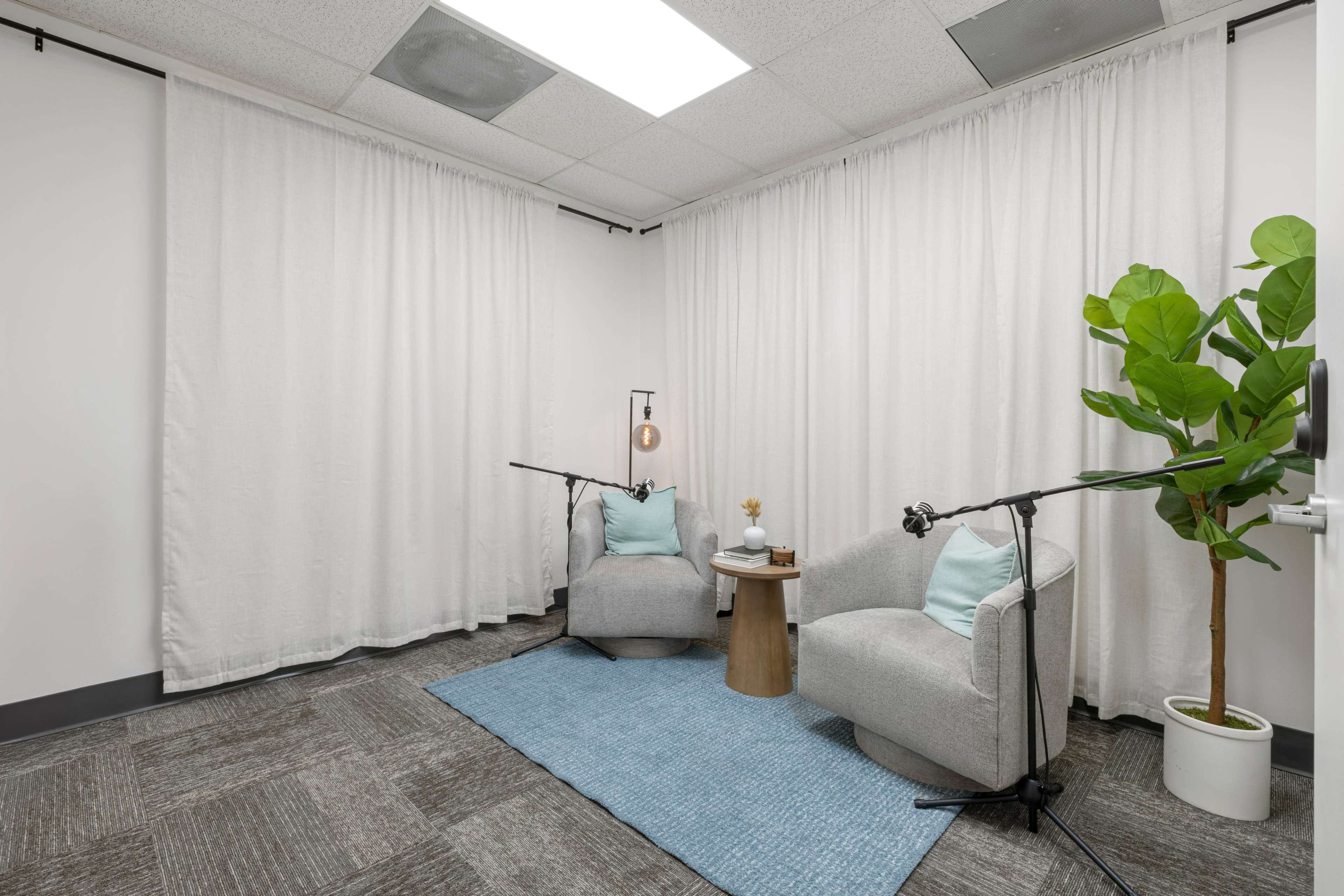 The image shows a small, modern conversation area with two gray armchairs, a round wooden table, a plant, and sound equipment positioned around a blue rug.